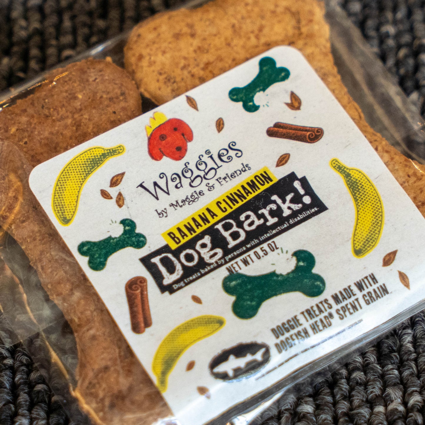 A close-up of packaged dog treats showing two brown bone-shaped treats in a clear plastic bag with a white label reading 'Wagges by Maggie & Friends Dog Bark! Banana Cinnamon' and illustrations of bananas, bones, and cinnamon sticks.