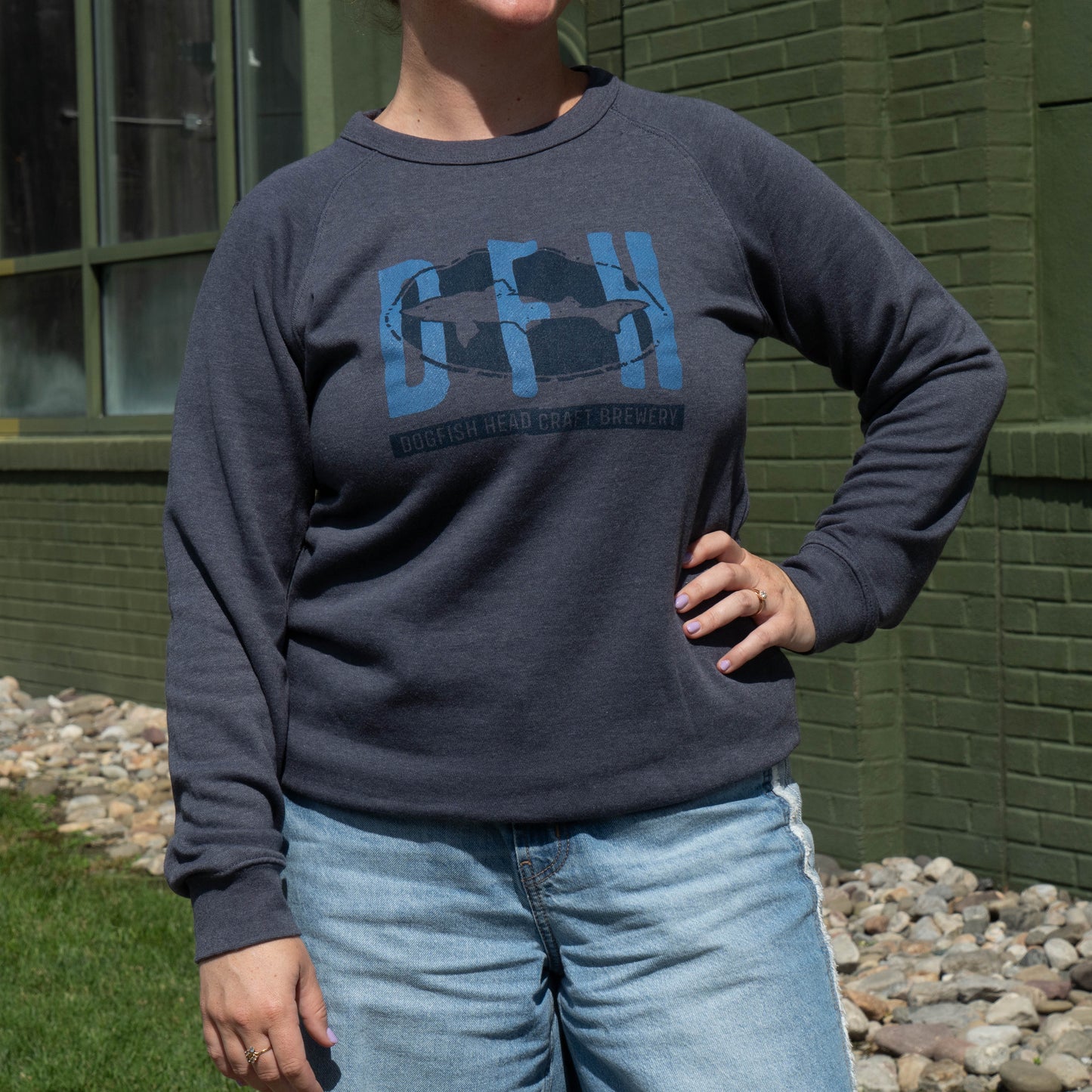 A person wearing a dark navy crewneck sweatshirt with a blue and black graphic on the chest, paired with light blue jeans, standing outdoors in front of a green brick building.