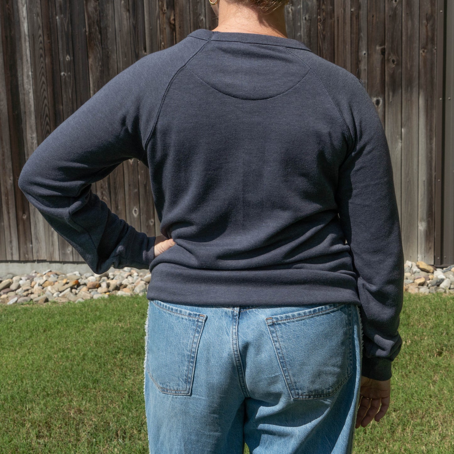 A person viewed from behind wearing a dark charcoal gray long-sleeve raglan sweatshirt and light blue jeans, standing outdoors in front of a wooden fence and grass.