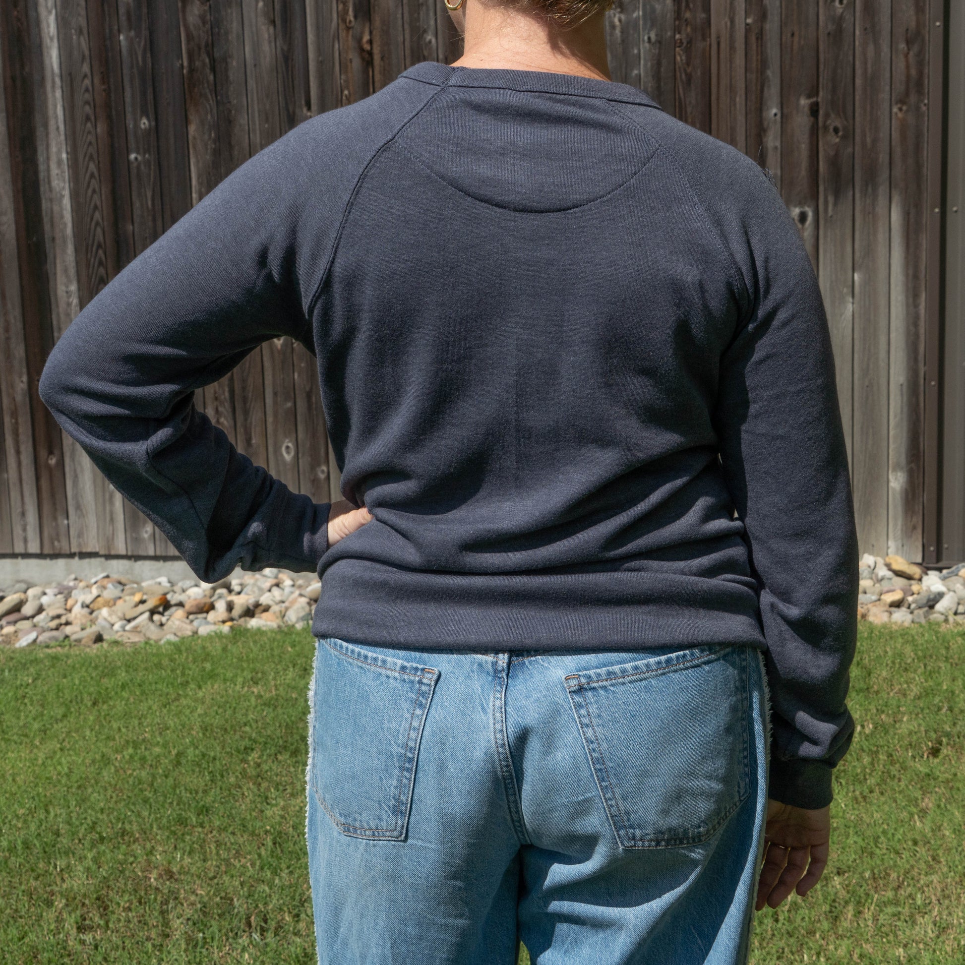 A person viewed from behind wearing a dark charcoal gray long-sleeve raglan sweatshirt and light blue jeans, standing outdoors in front of a wooden fence and grass.