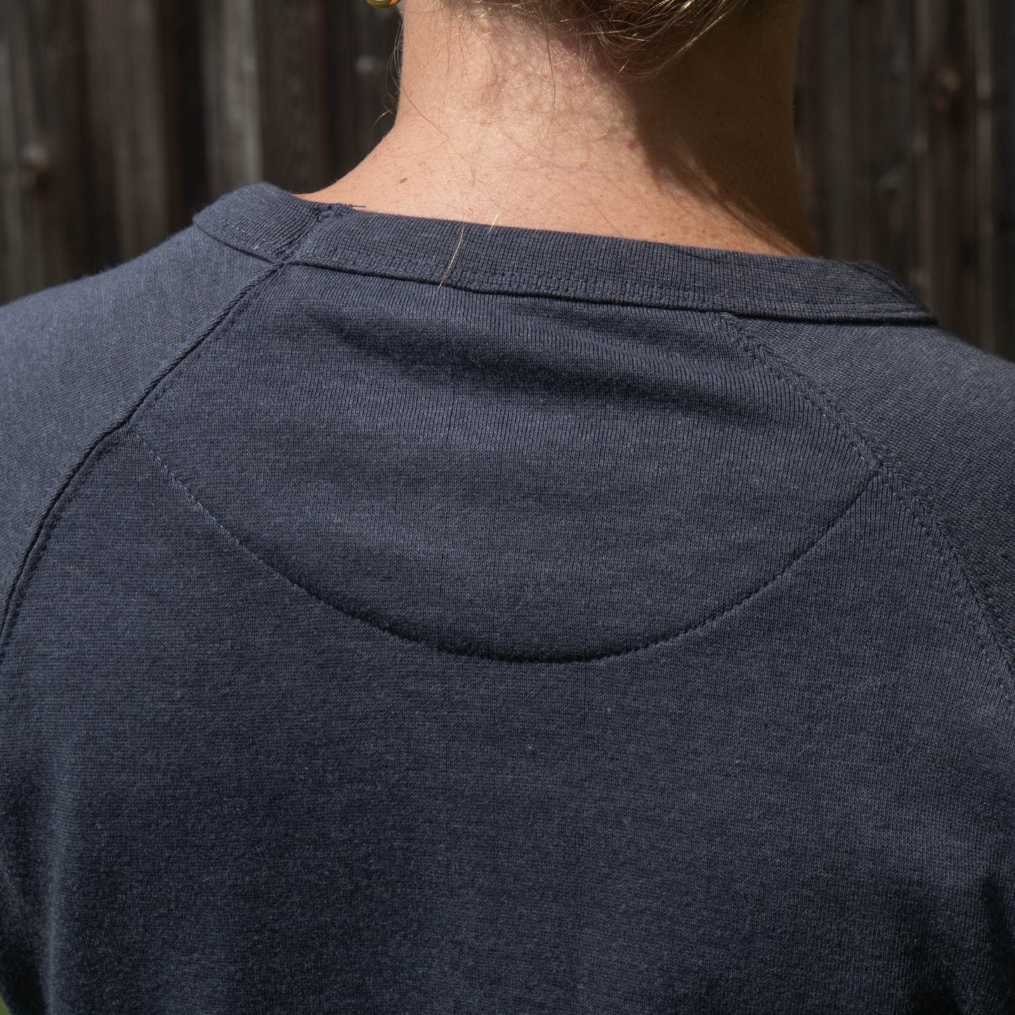Close-up back view of a person wearing a dark heathered navy raglan sweatshirt, showing the shoulder seam and collar stitching against a wooden fence background.