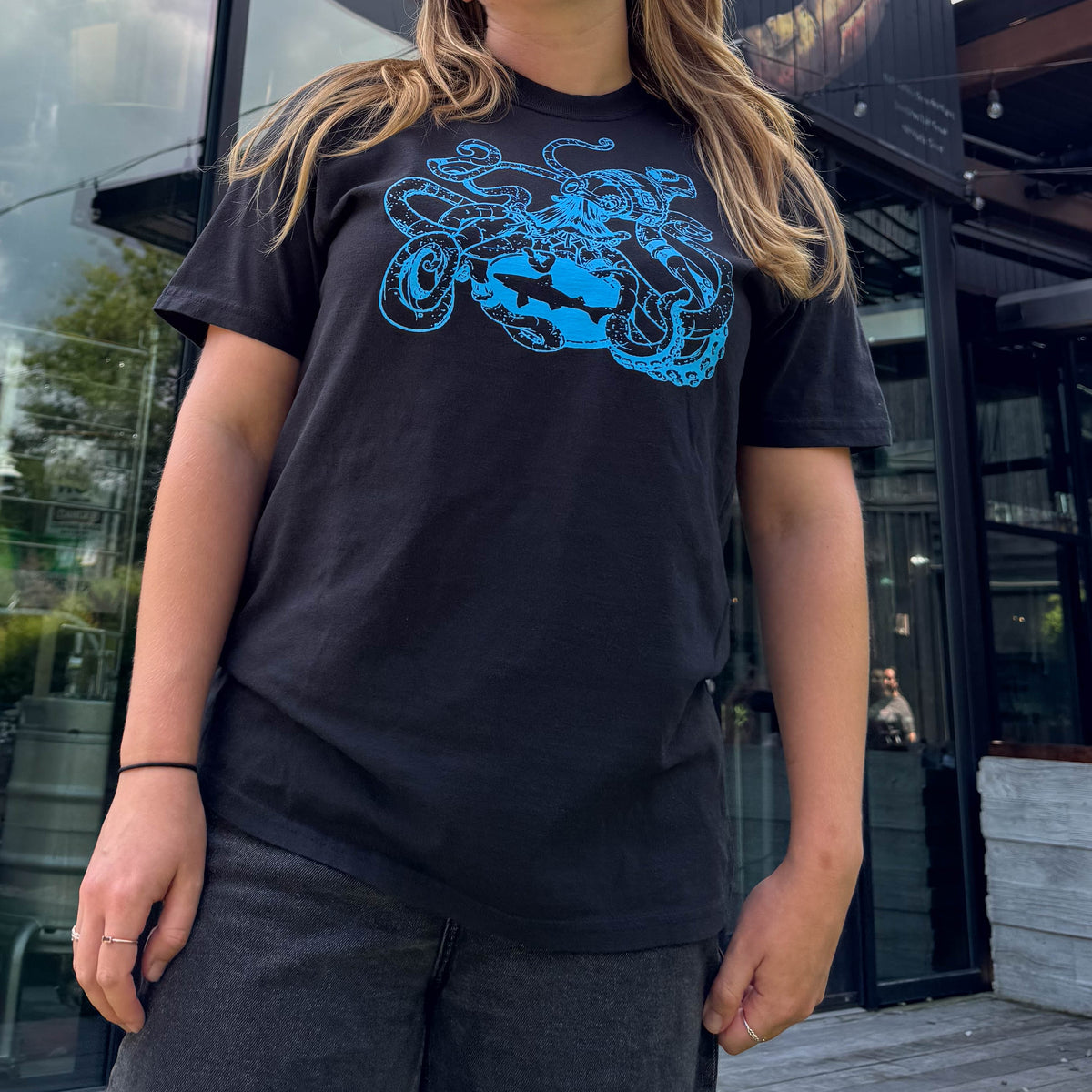 Black tee shirt with blue artwork of an octopus holding the dogfish head logo. On woman standing in front of building.