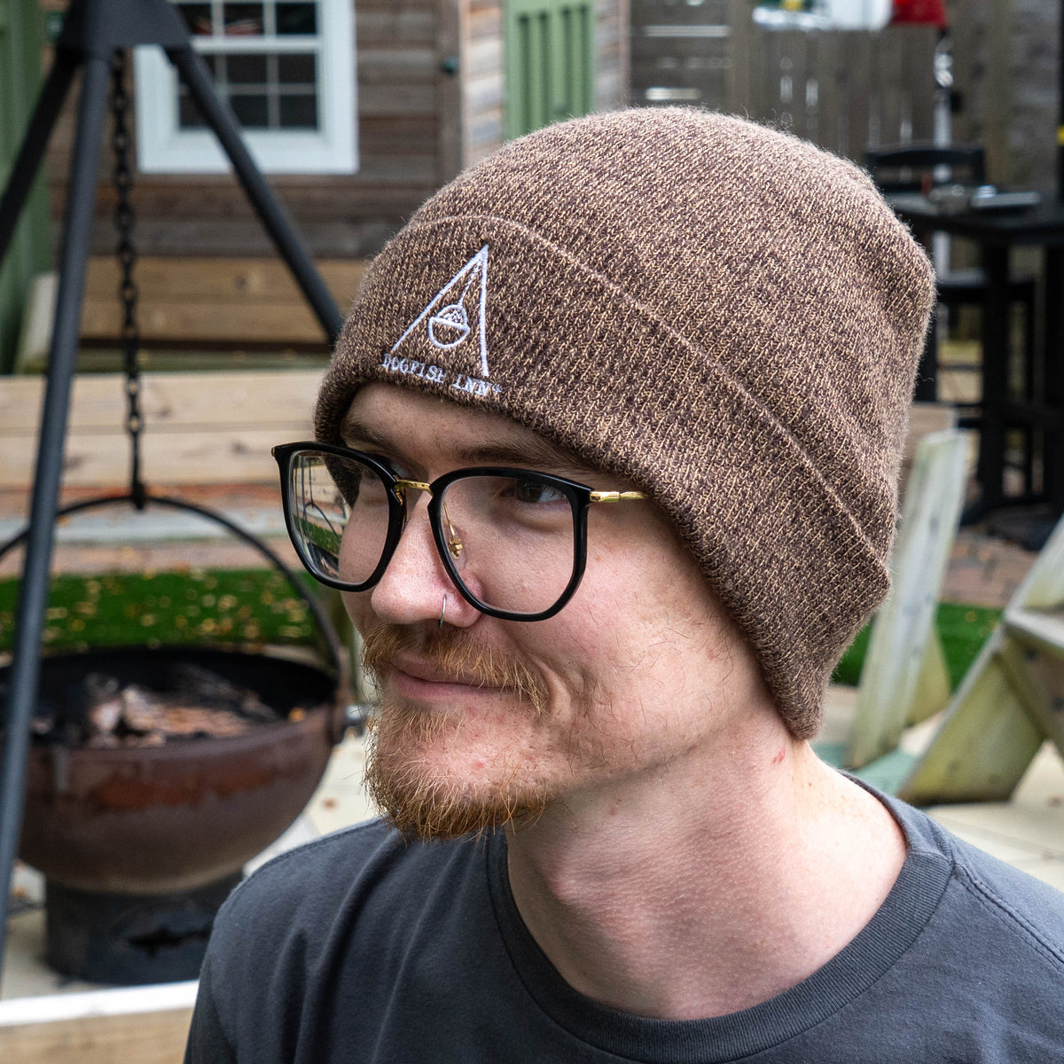 A person wearing an ARTEX KNITTING MILLS Brown Marled Firepit Beanie, a brown marled knit hat featuring a white embroidered logo of a fire pit inside a triangle.