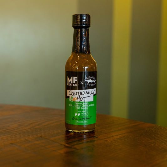 A bottle of Continually Hot Sauce by MF Heat Company sits on a wooden table with a blurred green and yellow background. The black and green label with white and red text highlights this fiery chimichurri hot sauce.