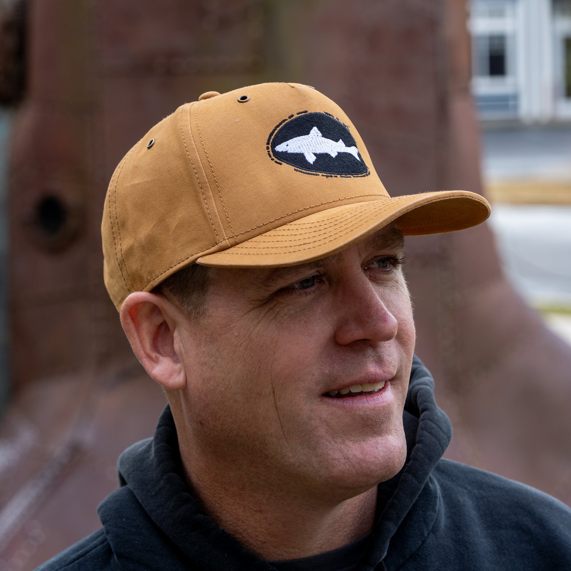 A man wears the Mirror Image Inc. Brown Richardson Five Panel Snapback with shark embroidery and a black hoodie while standing outdoors, smiling slightly in front of a blurred background.