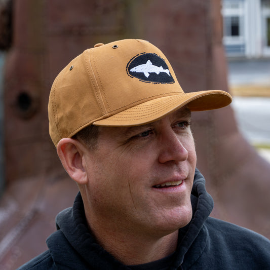 A man wears the Mirror Image Inc. Brown Richardson Five Panel Snapback with shark embroidery and a black hoodie while standing outdoors, smiling slightly in front of a blurred background.