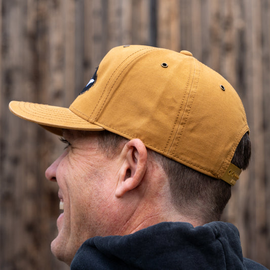 A man in profile, facing left, wears the Mirror Image Inc. Brown Richardson Five Panel Snapback and a black jacket, standing against a wooden fence background.