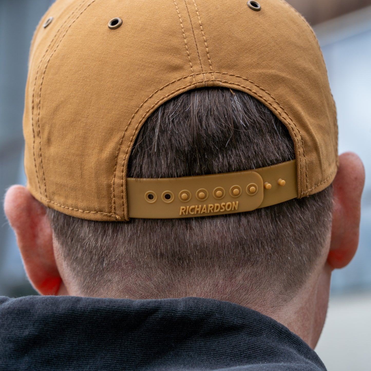 A close-up of the back of a person's head shows them in a dark top, wearing the Brown Richardson Five Panel Snapback by Mirror Image Inc., which features an adjustable strap with "RICHARDSON" and subtle shark embroidery.
