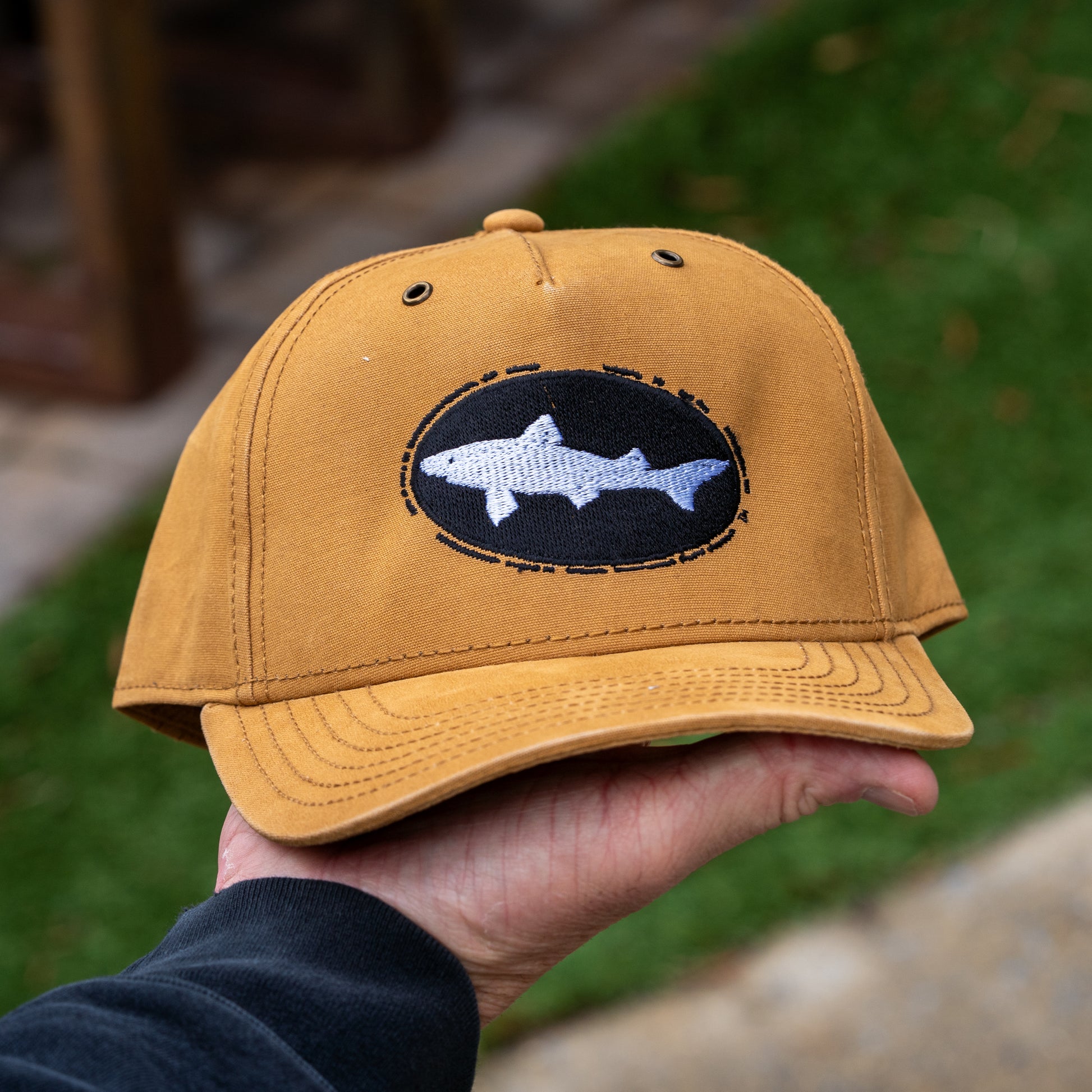 A Brown Richardson Five Panel Snapback by Mirror Image Inc. featuring an embroidered white shark patch inside a black oval is held outdoors, with grass and pavement blurred in the background.