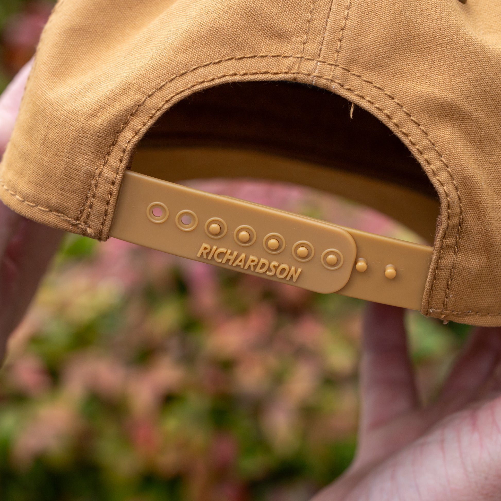 Close-up of the Brown Richardson Five Panel Snapback by Mirror Image Inc., held in hands, highlighting its adjustable plastic strap embossed with "RICHARDSON." Blurred foliage forms the background.