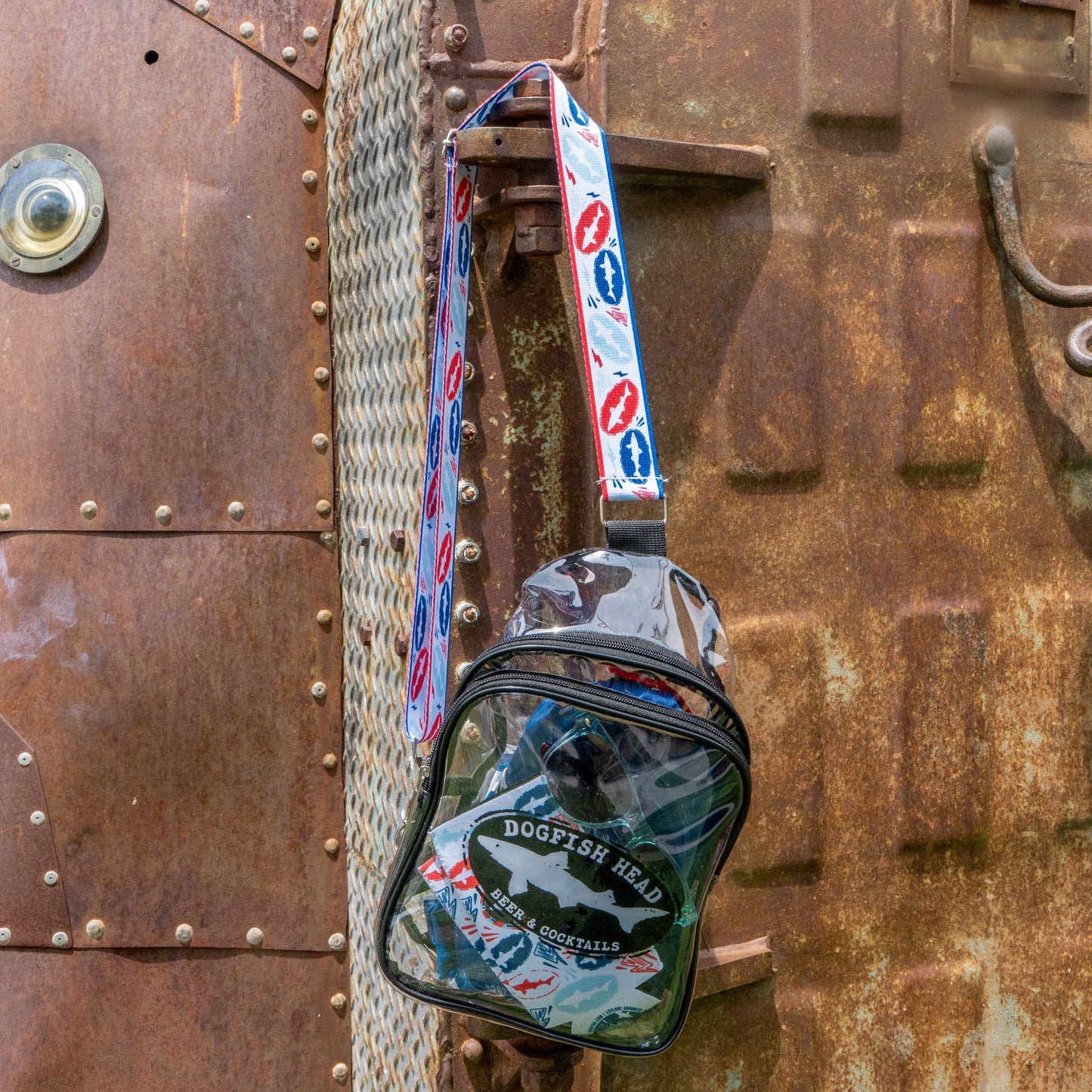 A clear Merchtank Drunk Summer Louder Festival Kit bag with a patterned strap hangs on a rusty metal surface with bolts, a circular window, and textured rope—your perfect summer festival essential. Drink Summer Louder!.
