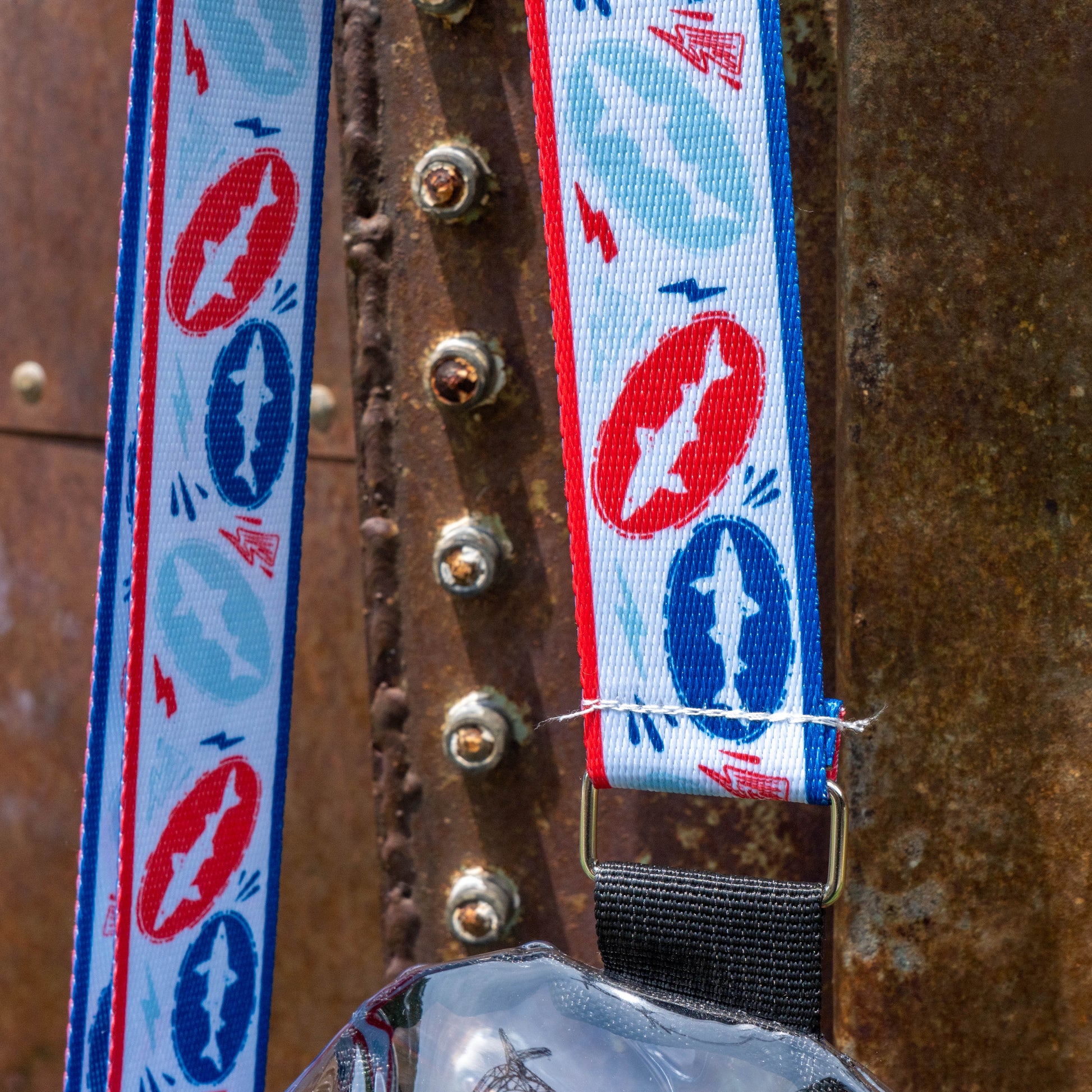 A close-up of the Drunk Summer Louder Festival Kit lanyard by Merchtank, featuring a red, white, and blue airplane pattern, hanging from a rusty metal surface—perfect for making summer festivals unforgettable.