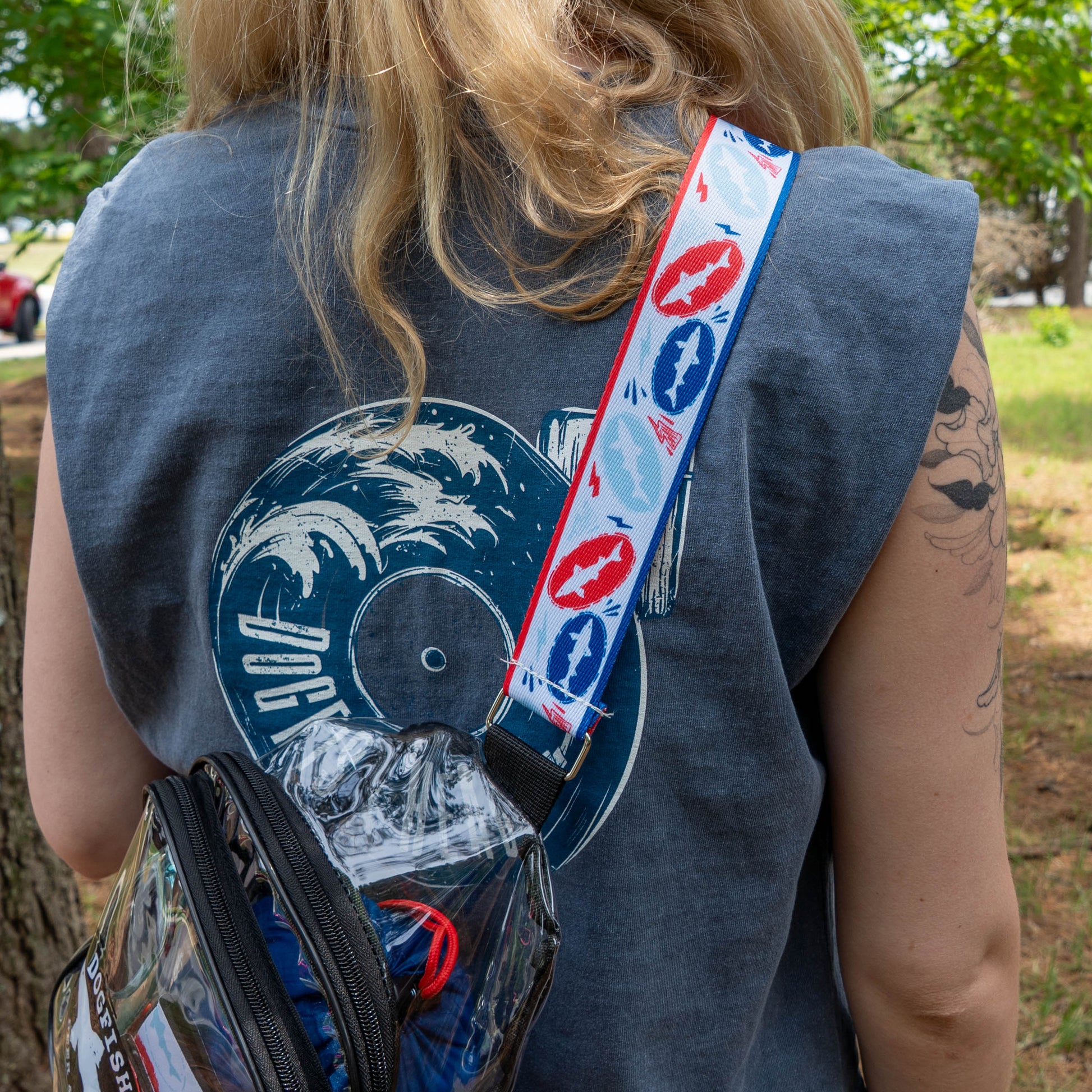 Wearing the Merchtank Drunk Summer Louder Festival Kit, featuring a sleeveless wave-and-vinyl shirt, clear crossbody bag, and colorful strap, a blonde-haired person embodies true festival style amid lush greenery.