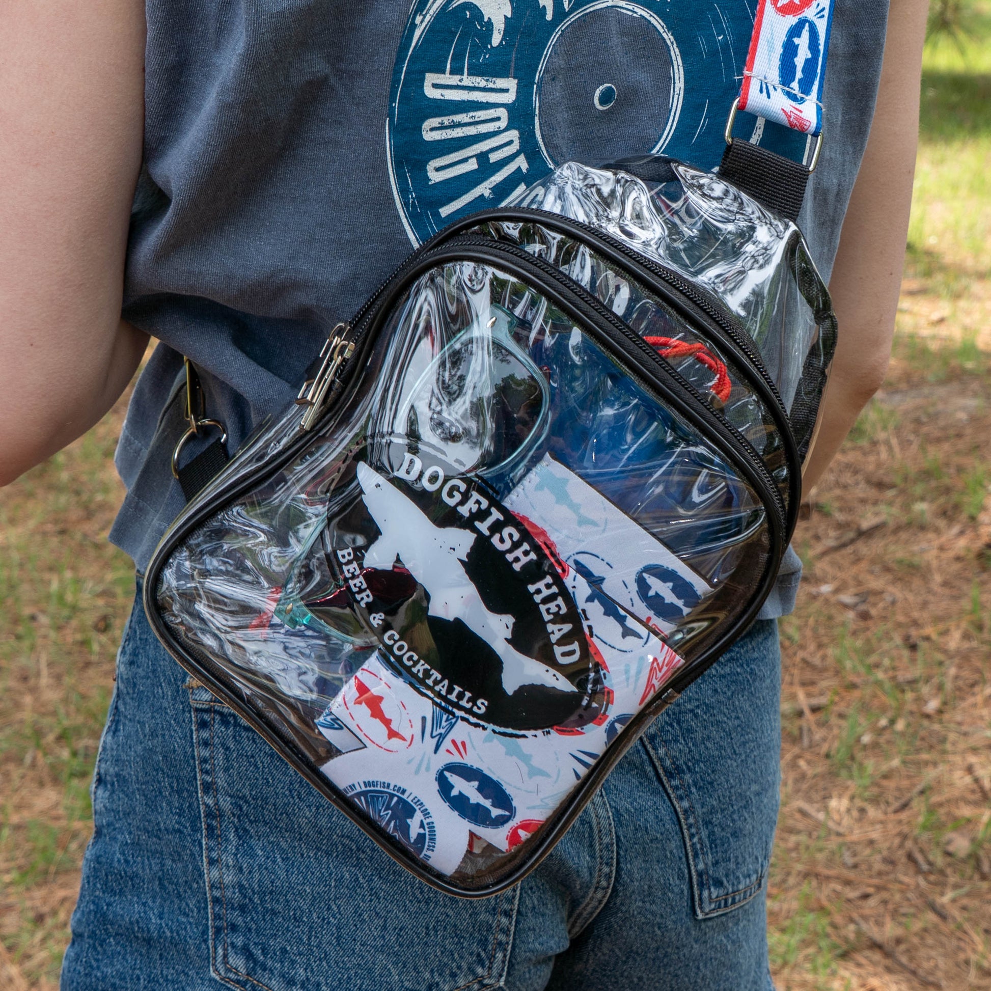 Wearing denim jeans and a blue Merchtank t-shirt, a person carries a clear crossbody bag filled with Drunk Summer Louder Festival Kit items and stickers—summer festival essentials—while standing outdoors on grass and pine needles.