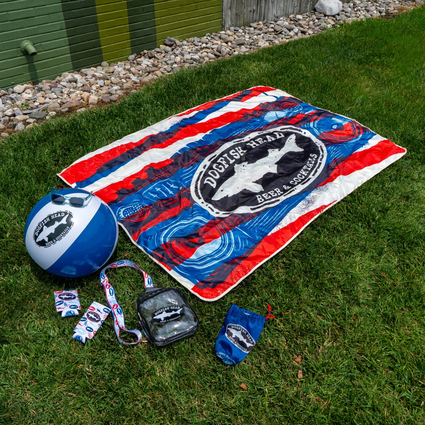 The Merchtank Drunk Summer Louder Festival Kit on grass features summer essentials like a striped towel, blue-and-white beach ball, pins, lanyard, clear pouch, and drawstring bag—perfect for festivals with a vibrant backdrop of rocks and painted wall.