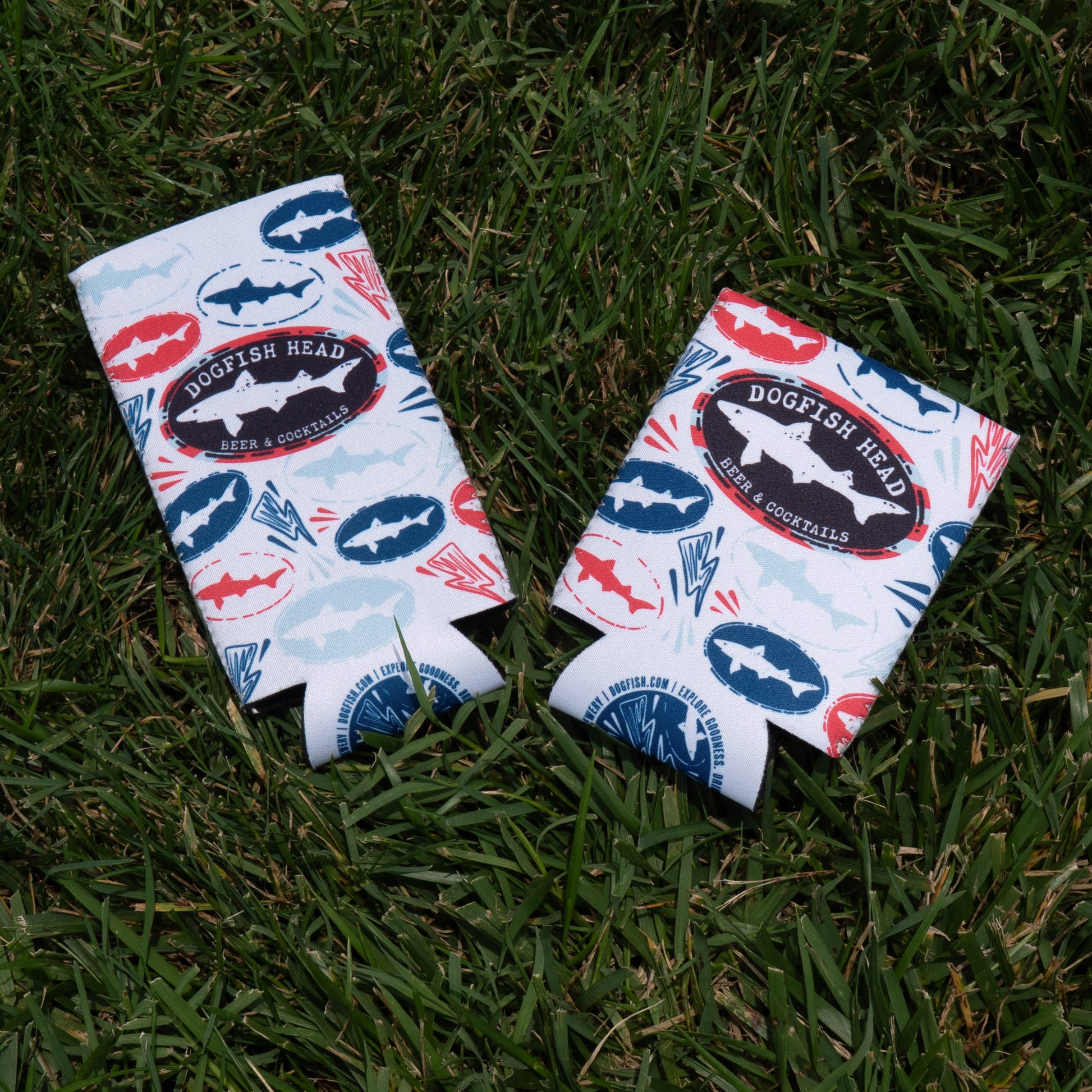 Two Merchtank Drunk Summer Louder Festival Kit beer can coolers with colorful shark patterns rest on green grass—must-haves for any summer festival. Keep your drinks cool and celebrate all season in style.