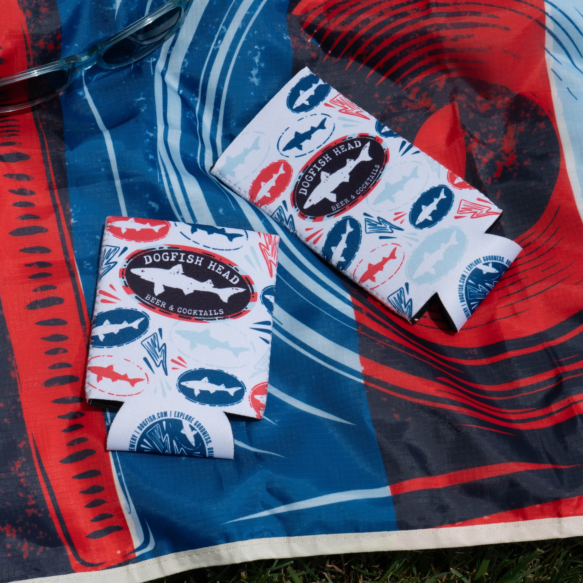 Two Merchtank Drunk Summer Louder Festival Kit can coolers with blue and red shark logos sit on a vibrant blue, red, and white cloth—perfect summer festival essentials. A pair of sunglasses is nearby. Drink Summer Louder!.