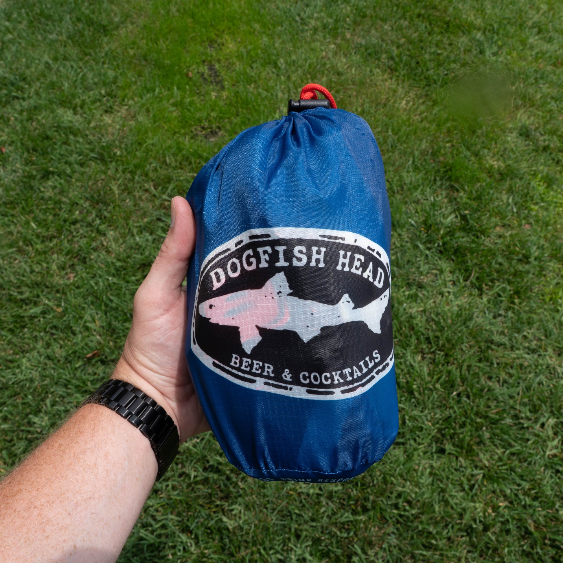 A hand holds a blue Drunk Summer Louder Festival Kit bag by Merchtank, featuring the brand’s logo—perfect for summer festival essentials—while standing on green grass and wearing a black wristwatch.