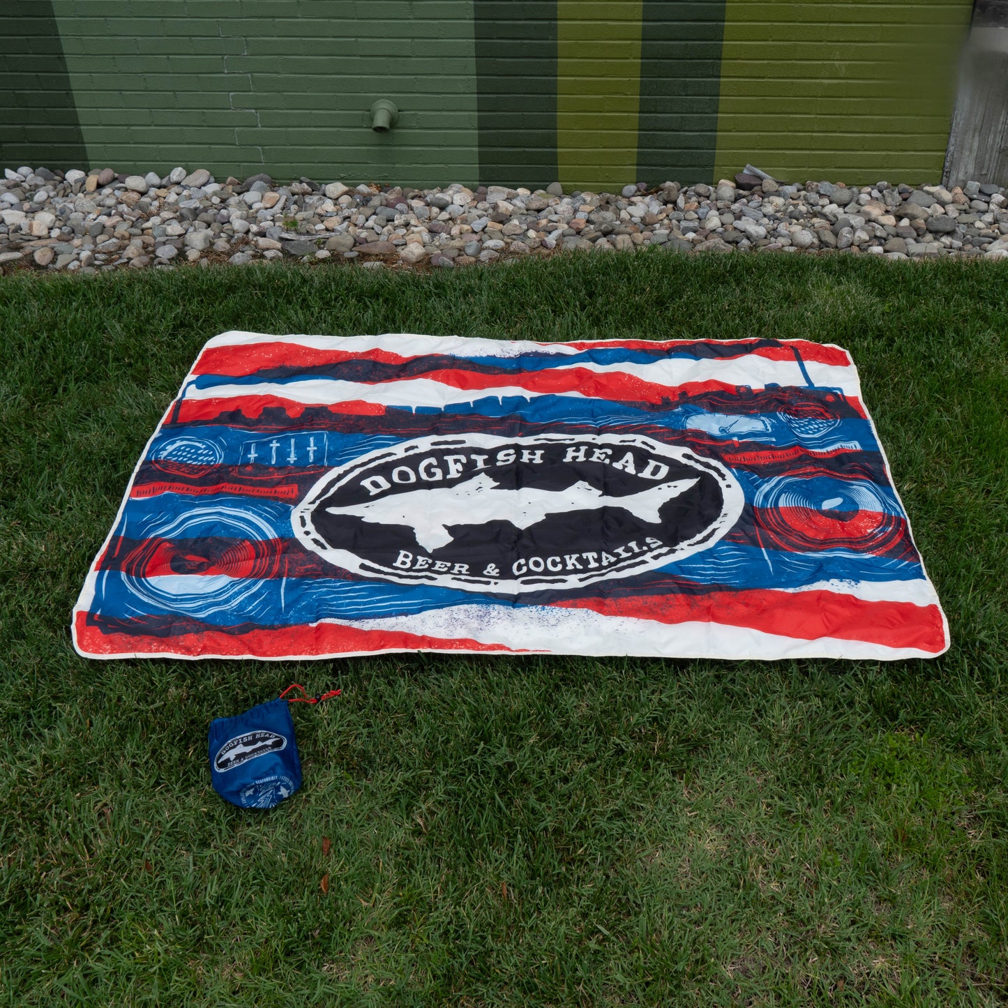 A red, white, and blue blanket from the Merchtank Drunk Summer Louder Festival Kit is spread on green grass near a brick wall. A matching blue carrying pouch lies nearby—ideal for your summer festival essentials.