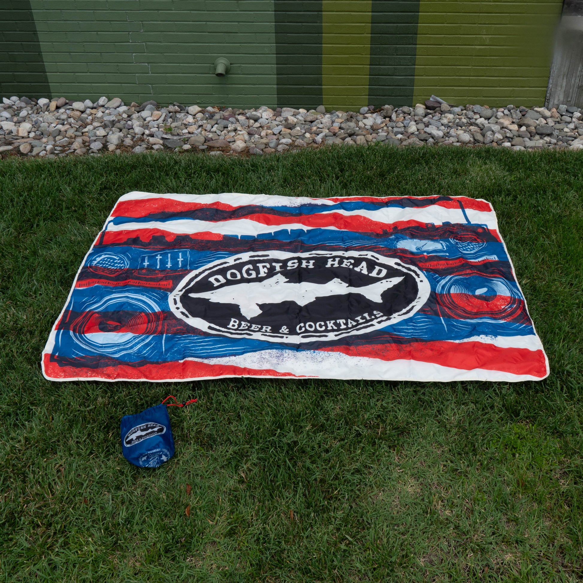 A red, white, and blue blanket from the Merchtank Drunk Summer Louder Festival Kit is spread on green grass near a brick wall. A matching blue carrying pouch lies nearby—ideal for your summer festival essentials.