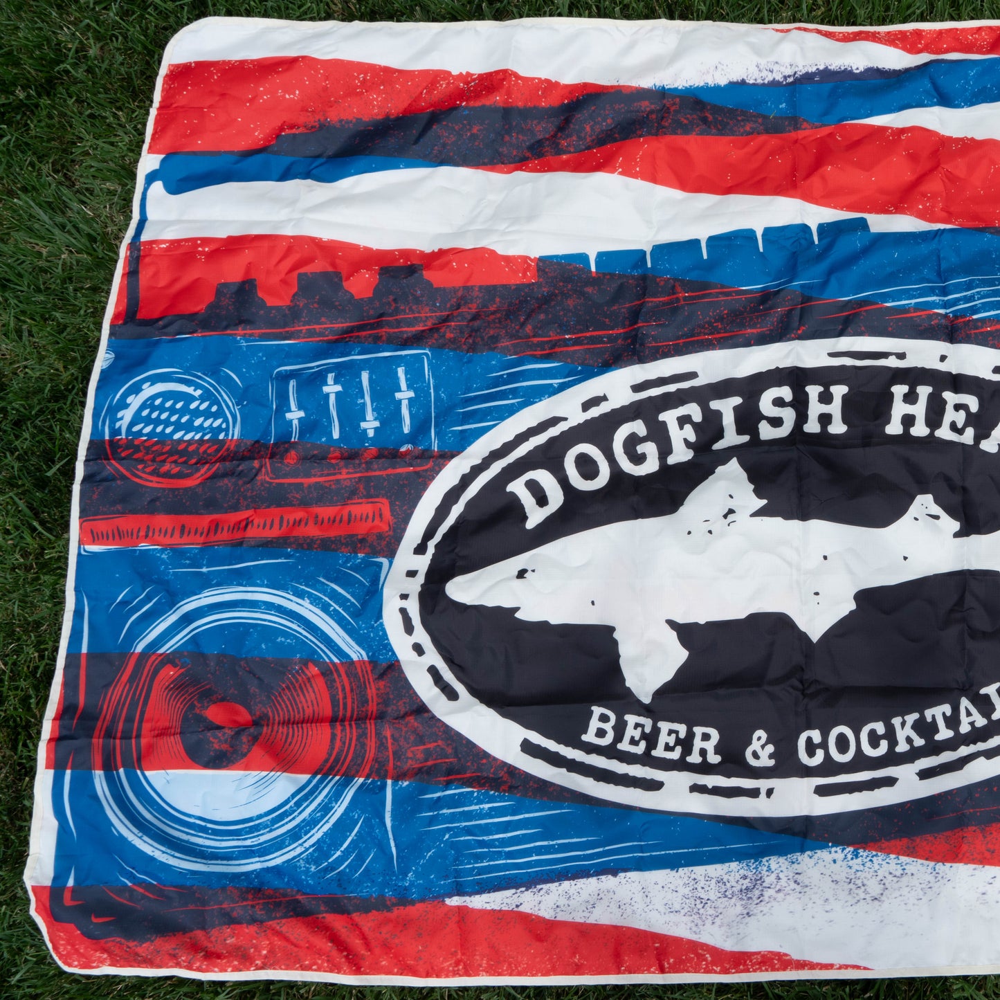 A vibrant blanket on grass displays the Merchtank Drunk Summer Louder Festival Kit logo with a white shark, bold text, red, white, and blue designs, and audio mixer art—must-haves for summer festivals.