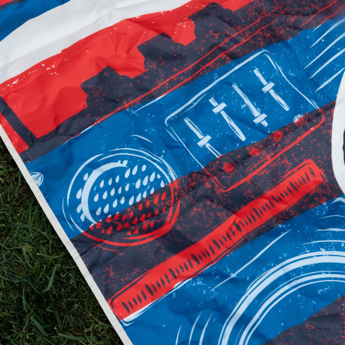 A close-up of a vibrant graphic on fabric, depicting a red, white, and blue boombox—part of the Merchtank Drunk Summer Louder Festival Kit—showcasing speakers and equalizer details as it rests on green grass. Summer vibes amplified!.