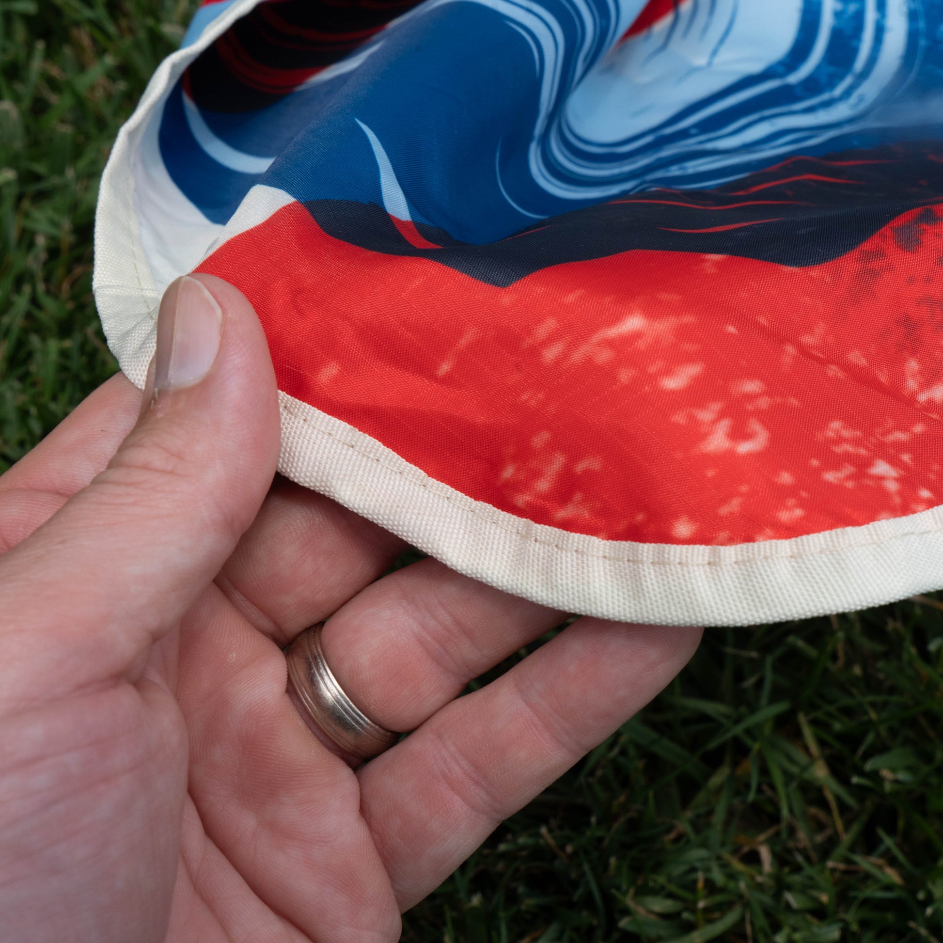 A hand wearing a silver wedding ring holds Merchtank's Drunk Summer Louder Festival Kit—colorful fabric with red, blue, and white patterns—above green grass, capturing perfect summer festival vibes.