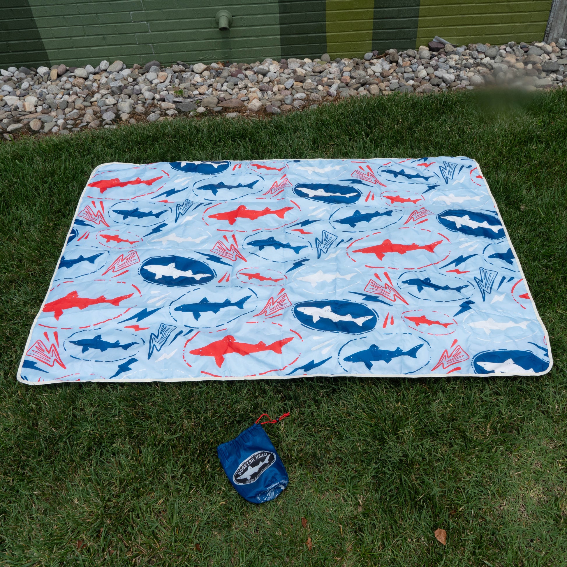 The Merchtank Drink Summer Louder Festival Kit features a rectangular picnic blanket with a blue, red, and white shark pattern on green grass. A blue carrying pouch is close by, with rocks and a green painted wall in the background.