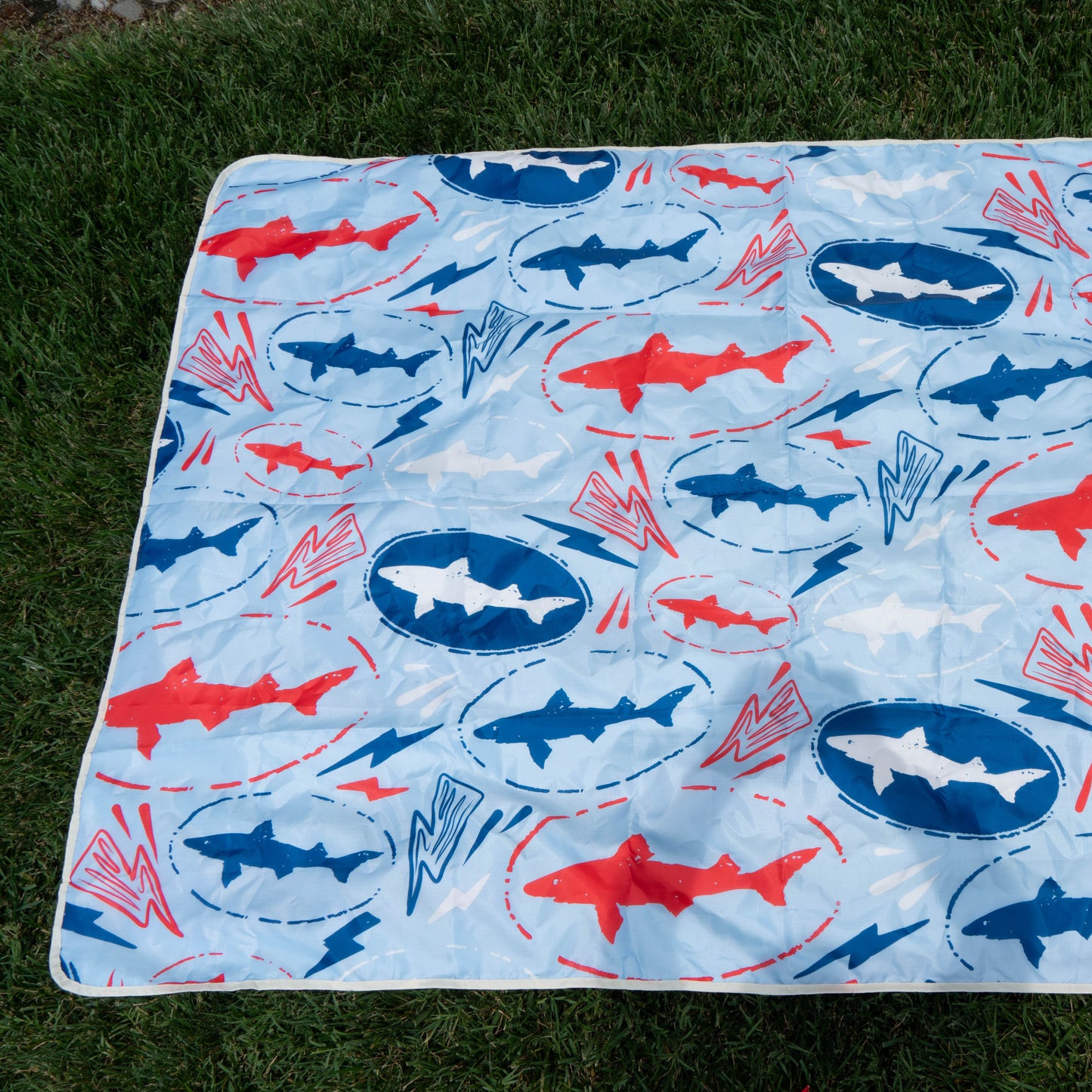 A crumpled picnic blanket with red and blue sharks, blue circles, and red zigzags lies on green grass—a must-have from the Merchtank Drink Summer Louder Festival Kit.