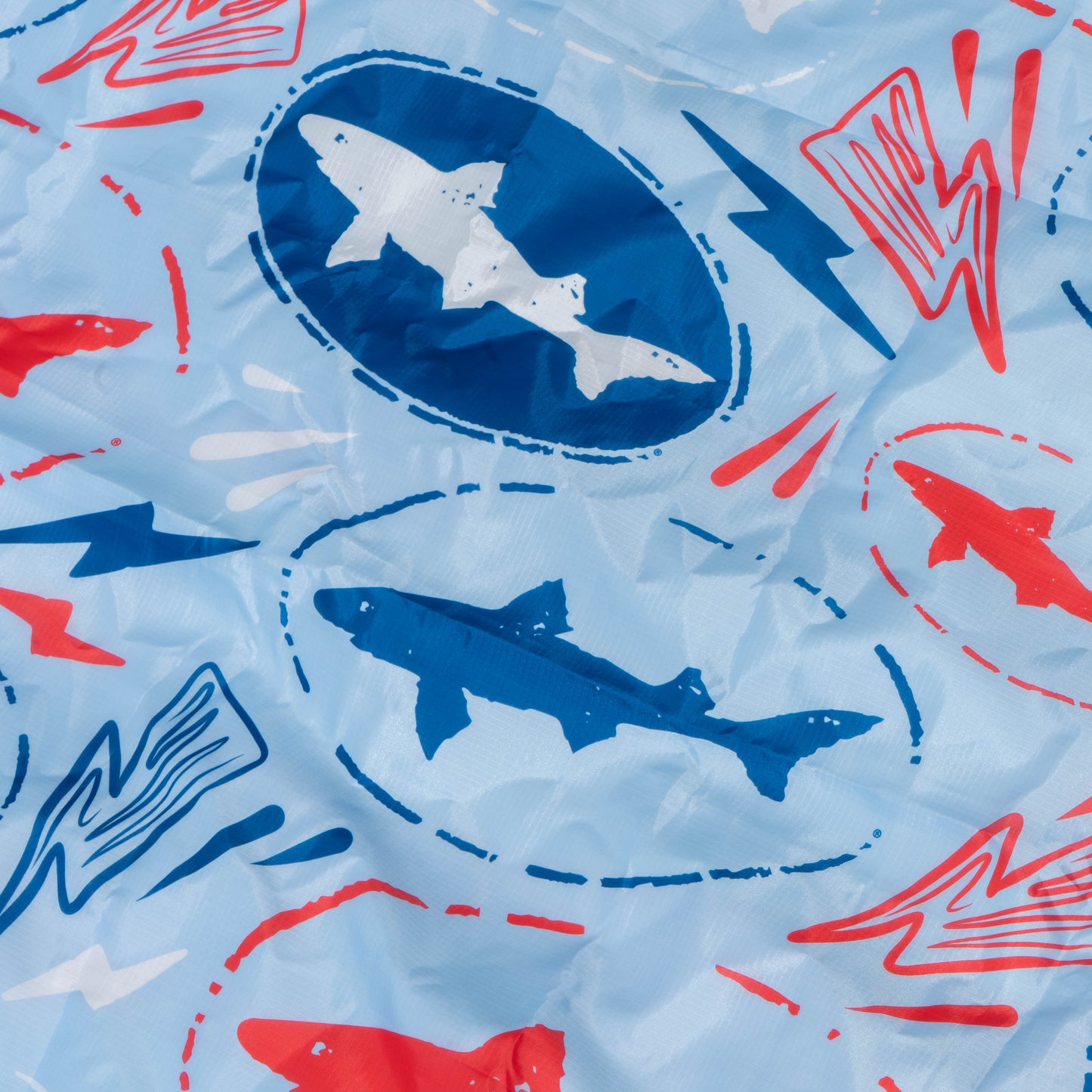 Light blue fabric with blue, white, and red sharks in ovals plus red and blue lightning bolts—great for summer crafts or customizing your Merchtank Drink Summer Louder Festival Kit. Fabric has a slightly wrinkled appearance.
