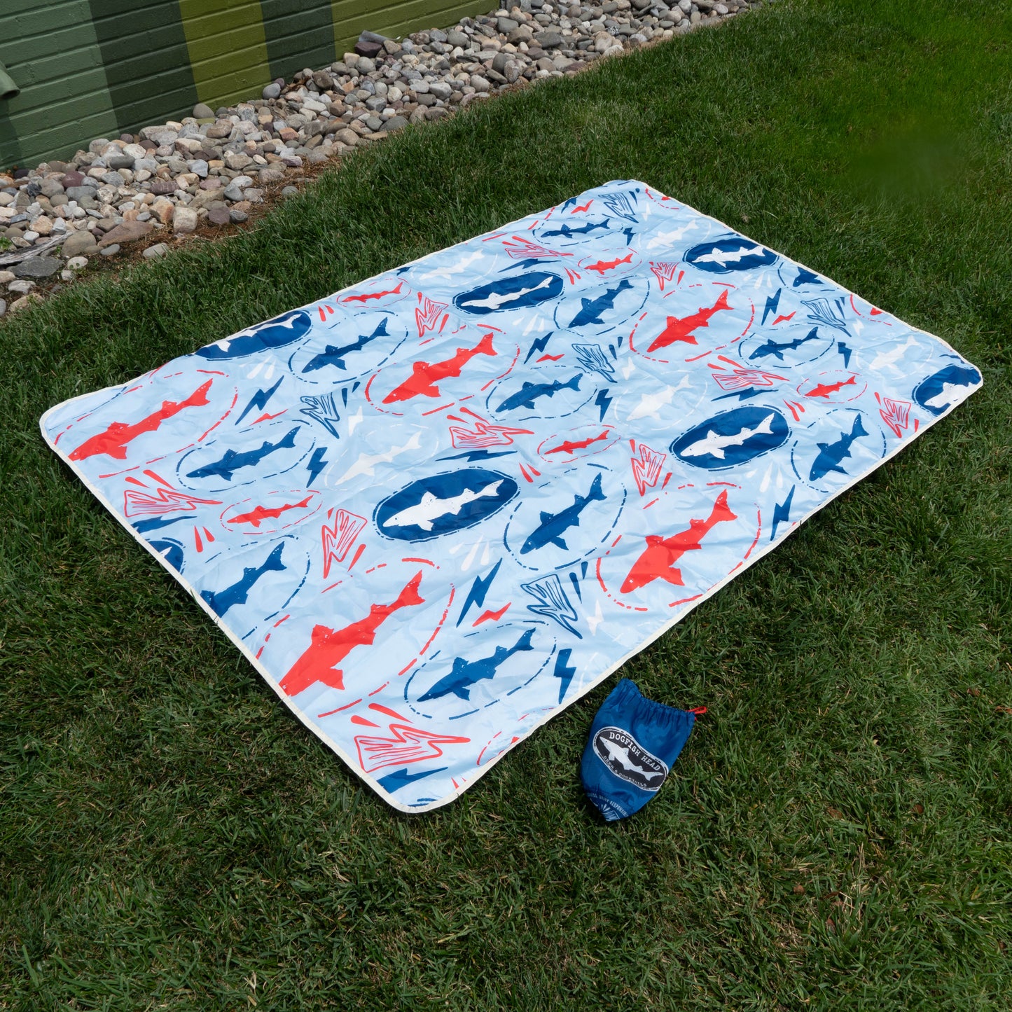 A light blue picnic blanket with red and blue shark patterns from the Drink Summer Louder Festival Kit by Merchtank is spread on grass, with a matching pouch nearby—ideal for festival essentials. Rocks and a building appear in the background.