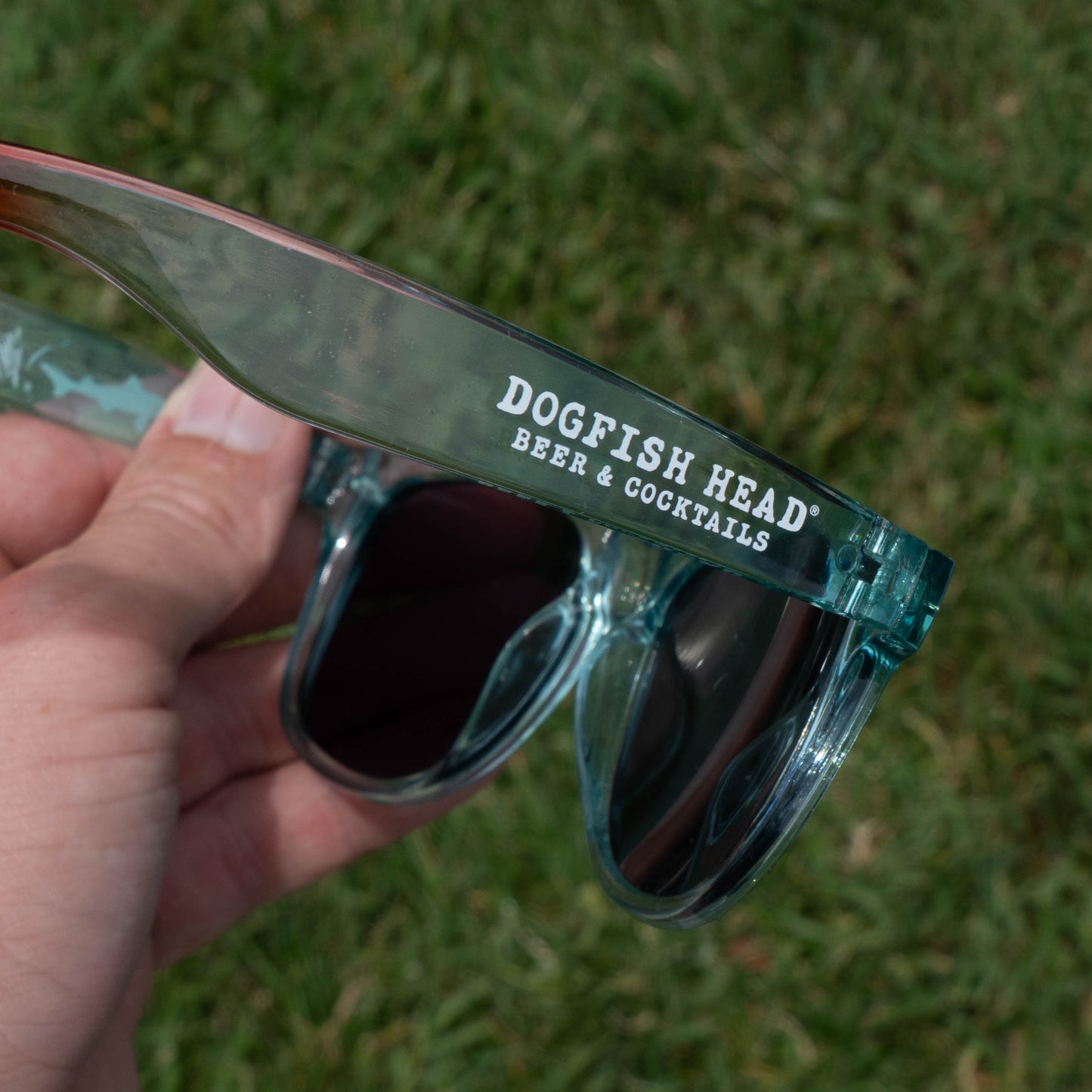 A hand holds a pair of teal sunglasses with "Dogfish Head Beer & Cocktails" printed in white—summer essentials from the Drink Summer Louder Festival Kit by Merchtank—against a blurred green grass background.