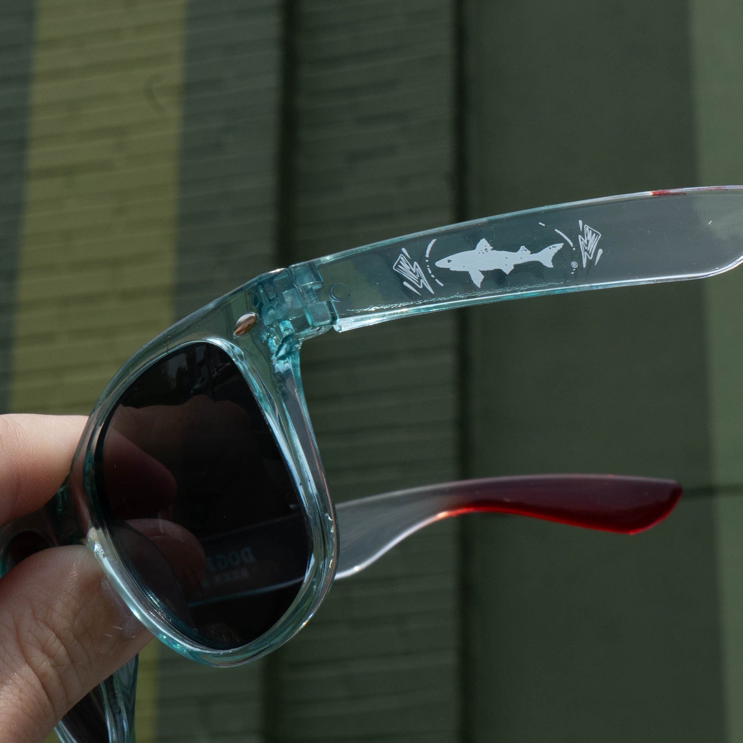 A hand holds Merchtank sunglasses with translucent blue frames, red arms, and a white shark graphic—fun festival essentials from the Drink Summer Louder Festival Kit. A green brick wall in the background sets the playful mood.
