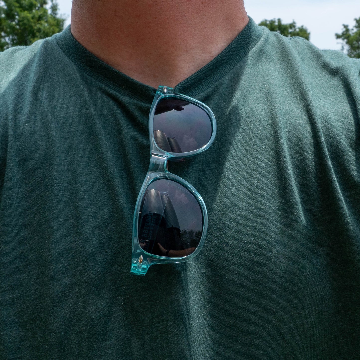 Wearing a green t-shirt with blue-rimmed sunglasses reflecting outdoor scenes, this person embodies festival vibes. Their face is hidden, but the Drink Summer Louder Festival Kit from Merchtank captures pure summer spirit.