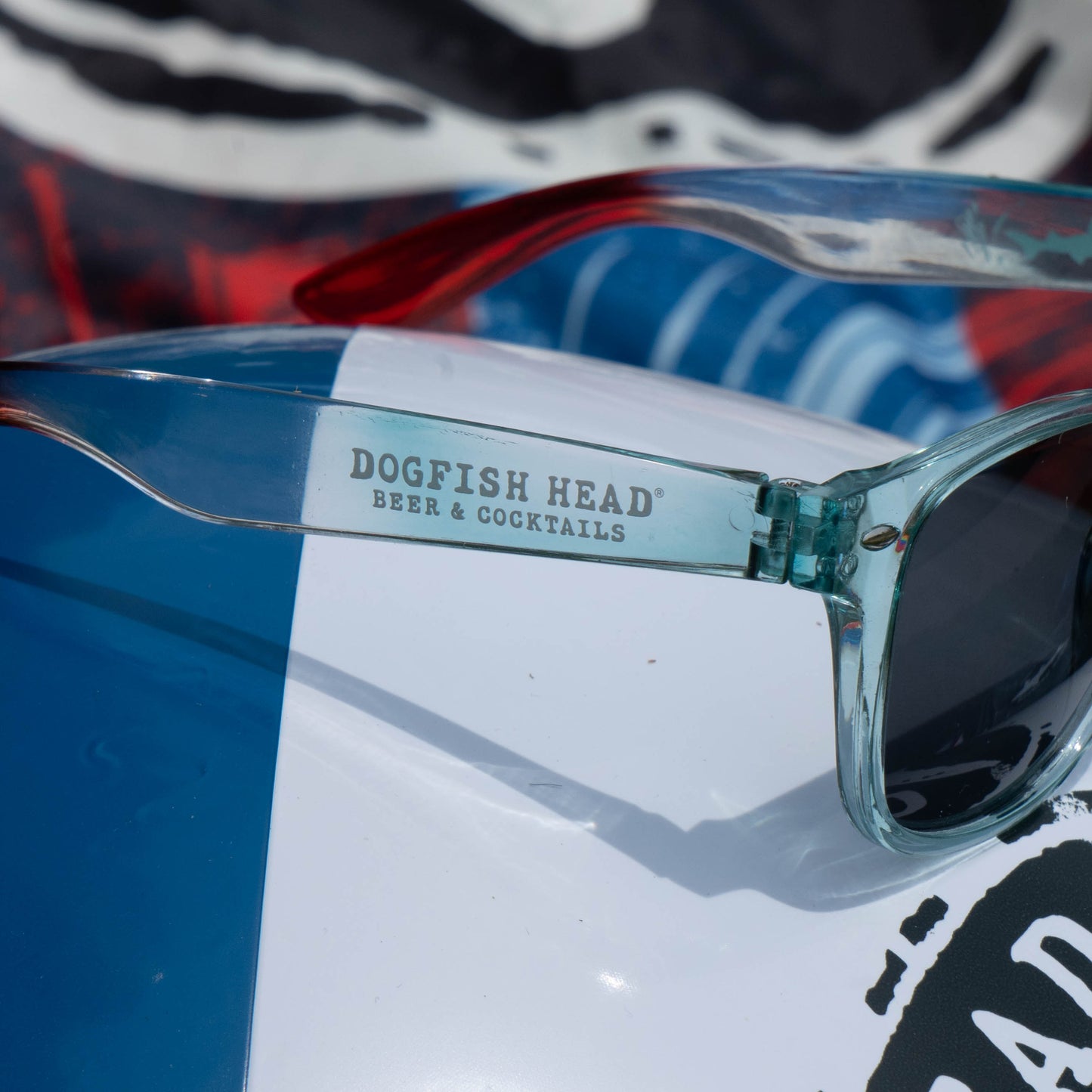 Close-up of transparent sunglasses with Dogfish Head Beer & Cocktails on the arm, resting on a colorful surface—capturing the Drink Summer Louder Festival Kit by Merchtank, perfect for summer events with upbeat, festive vibes.