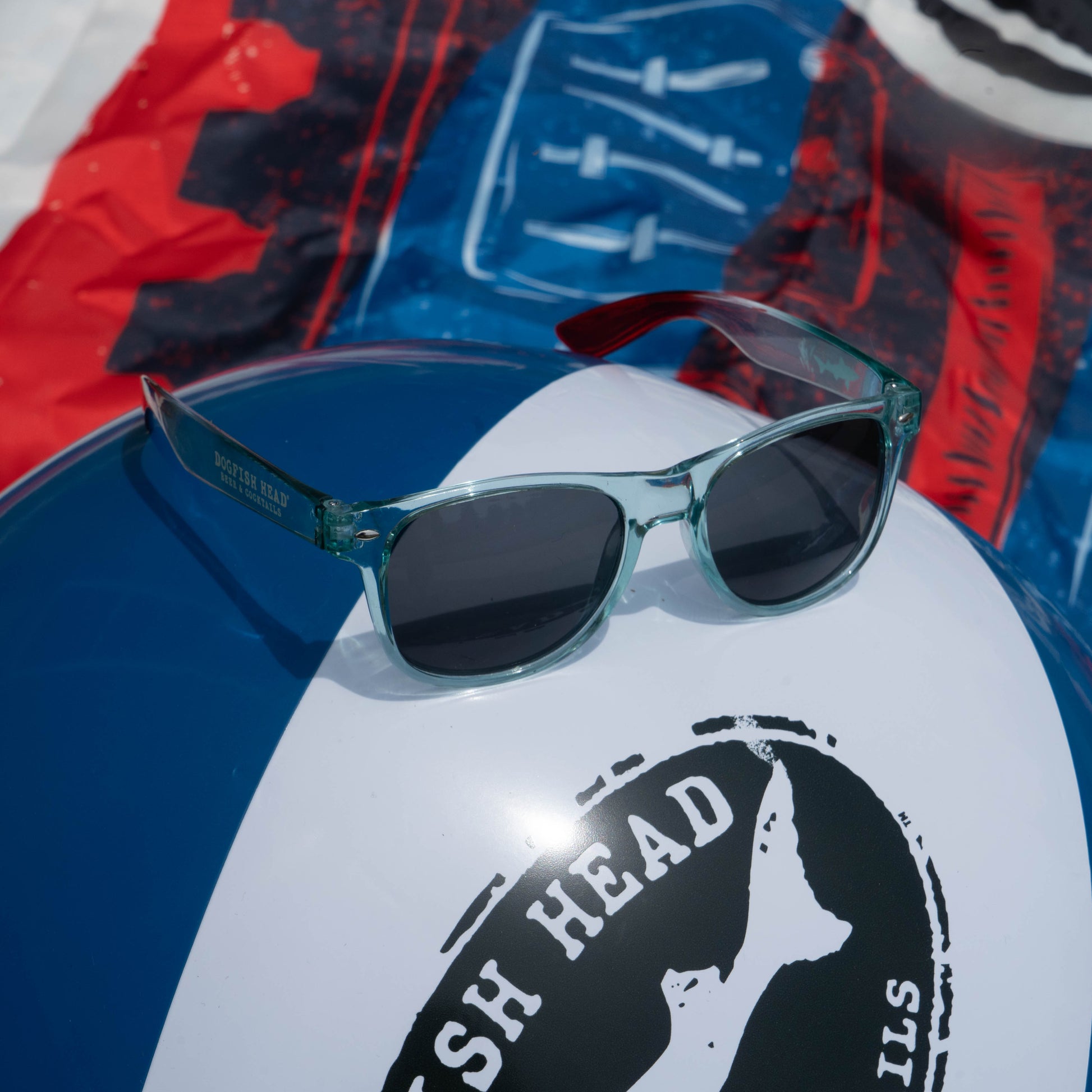 A pair of clear-framed sunglasses sits on a blue, white, and black beach ball featuring the Drink Summer Louder Festival Kit by Merchtank, with a colorful beach towel in the background—perfect summer festival essentials.