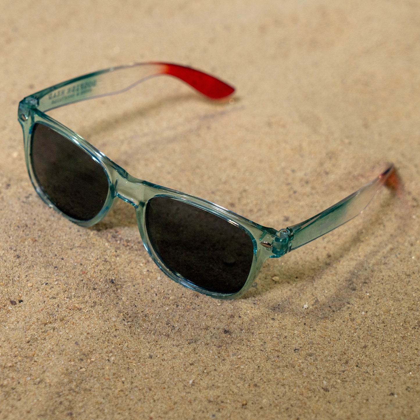 A pair of translucent teal sunglasses with dark lenses and red-tipped arms lies on sandy ground—true summer festival essentials from the Merchtank Drink Summer Louder Festival Kit.