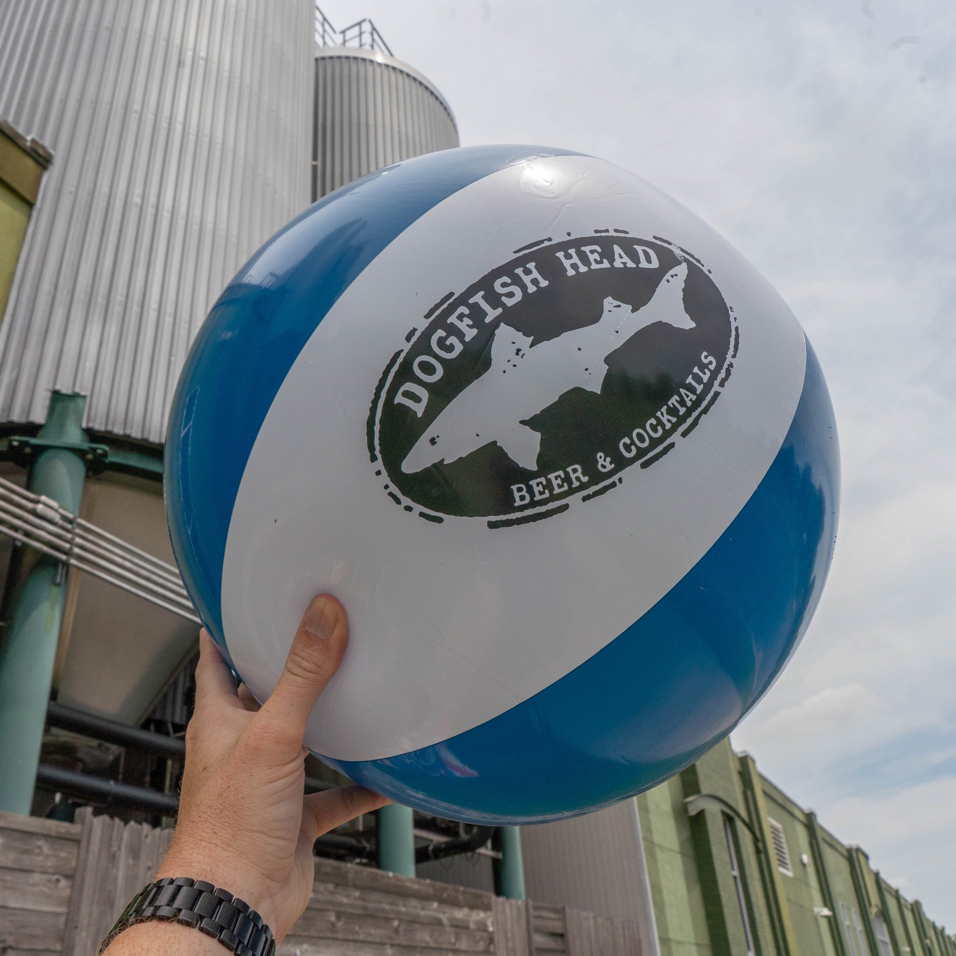 A hand holds up the Merchtank Drink Summer Louder Festival Kit beach ball—one of your summer festival essentials—in front of brewery tanks and a green building under a partly cloudy sky.