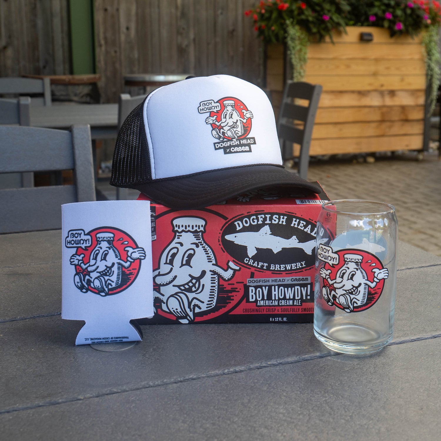 A Dogfish Head craft brewery trucker hat sits on a gray outdoor patio table, styled with a matching branded can cooler and a clear pint glass. The hat features a white front panel, black mesh back, and a cartoon dogfish logo with the text 'Dogfish Head Craft Brewery' and 'Boy Howdy!'.