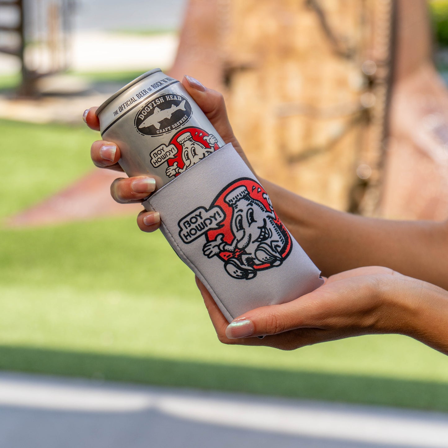 A person holds a silver can inside a white neoprene can cooler outdoors. The can cooler has red and black graphics and the text 'BOY HOWDY!' and Dogfish Head Craft Brewery logo