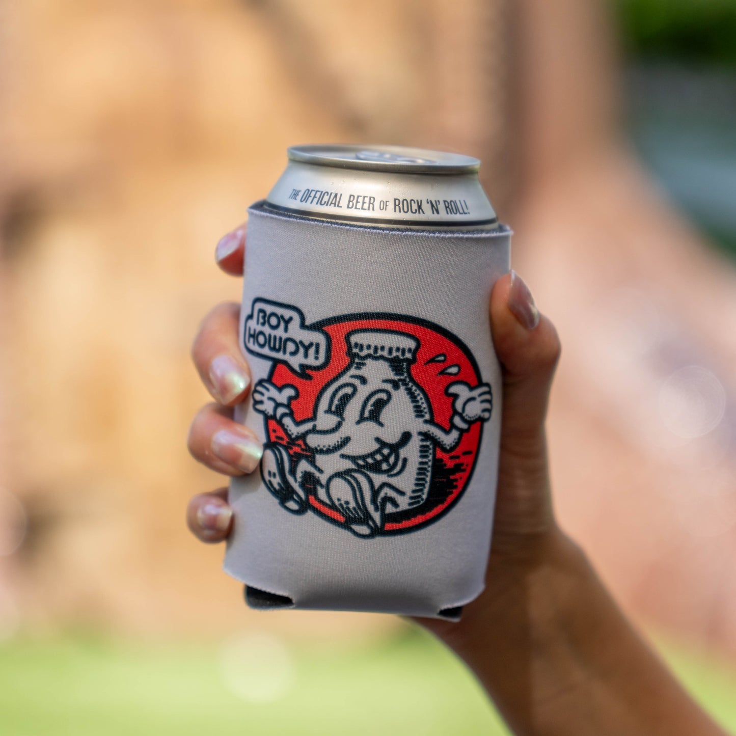 A hand holding a gray neoprene can cooler with a red and black cartoon graphic and the text 'BOY HOWDY!'. The can inside has 'OFFICIAL BEER OF ROCK 'N' ROLL!' printed around the rim.