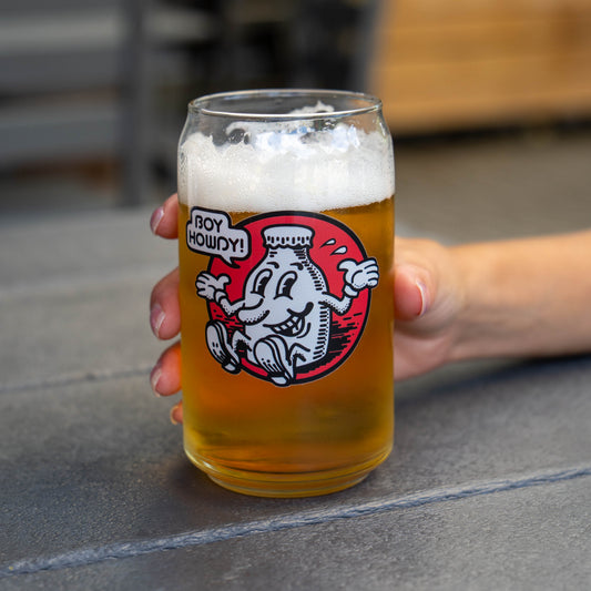A hand holds a clear beer glass filled with golden beer and white foam, featuring a graphic of a character with the text 'BOY HOWDY!' on a gray stone surface.