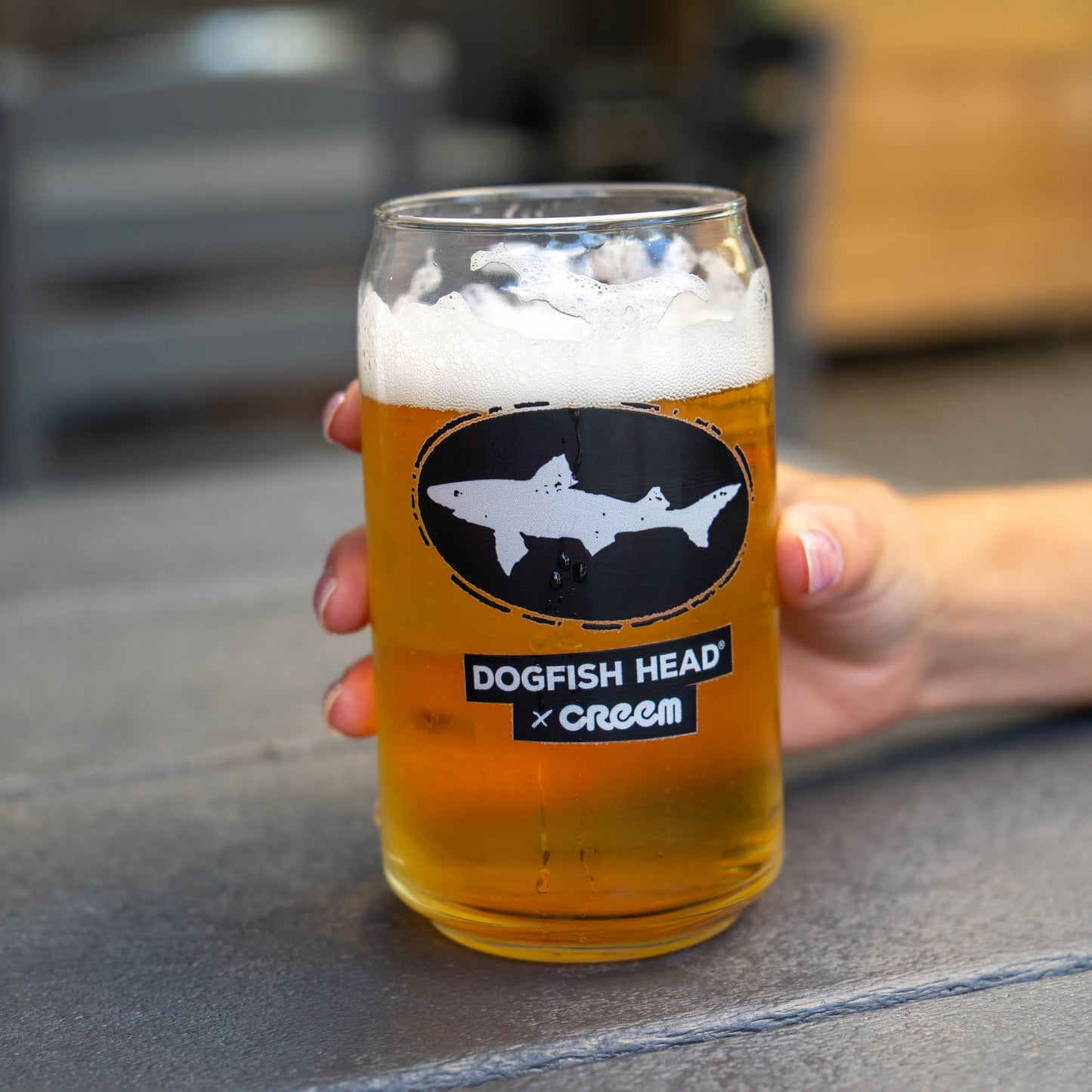 A hand holds a clear glass pint GLASS filled with golden beer and topped with white foam. The mug has a black oval shark logo and the text 'DOGFISH HEAD x CREEM' on the front, set against a blurred outdoor background.