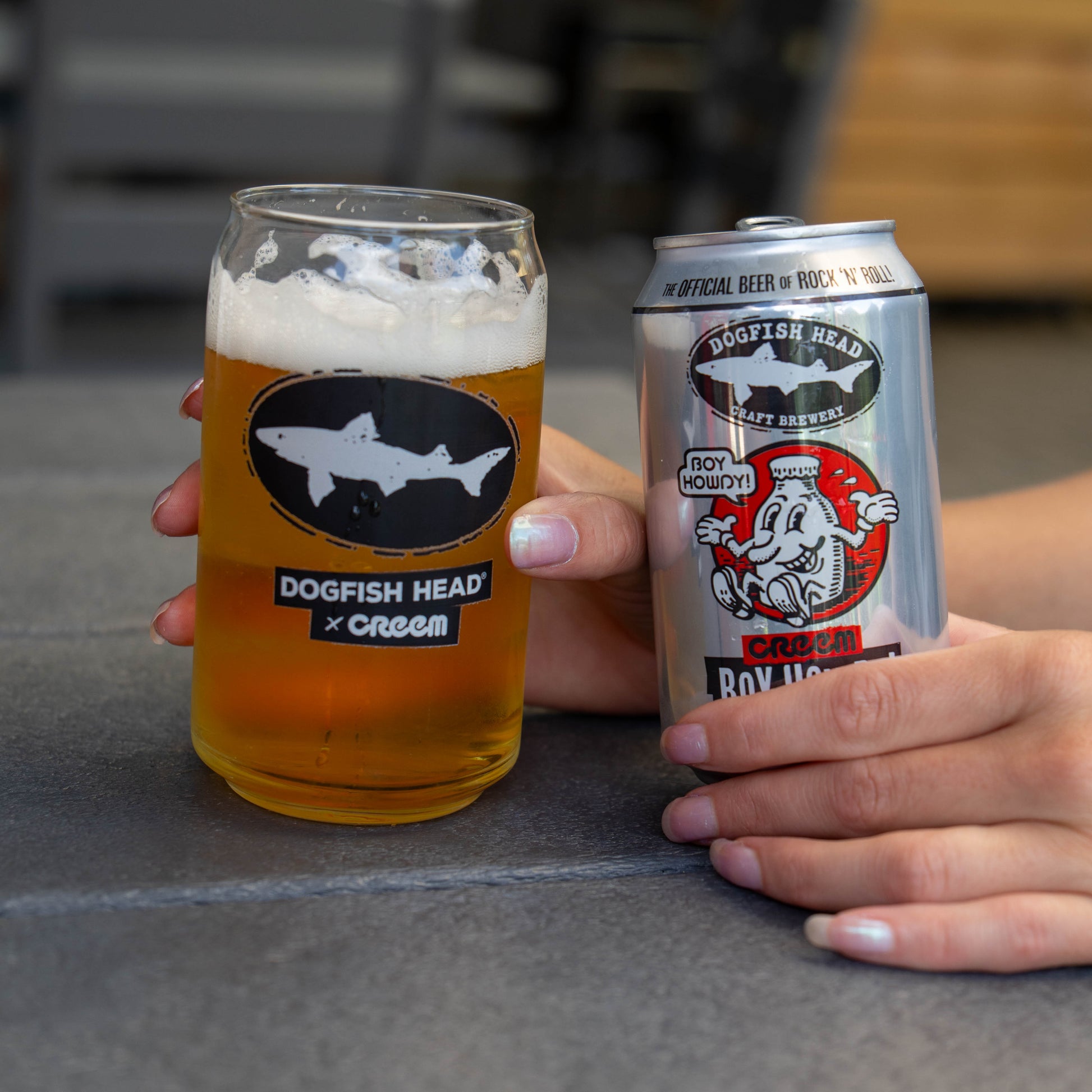 A hand holds a frosted glass filled with amber beer and white foam, printed with the Dogfish Head x Creem logo, while another hand holds a silver can of Dogfish Head Creem Boy Howdy beer next to it on a gray tabletop.