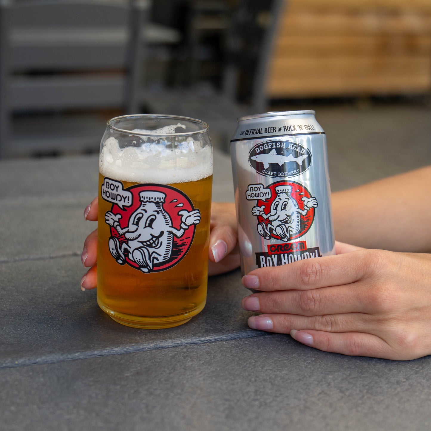 A clear pint glass and a silver aluminum can, both printed with the same graphic and the text 'Dogfish Head.' and 'Boy Howdy', held by hands over a gray stone surface.
