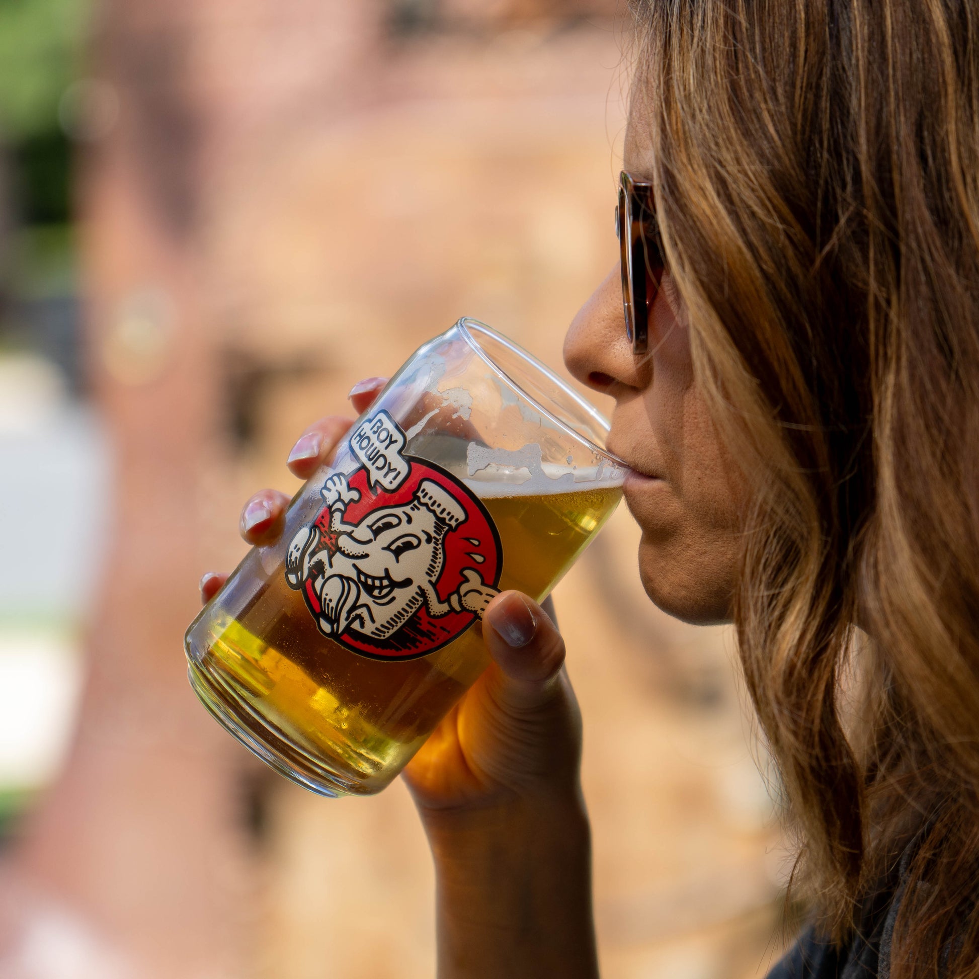 A close-up lifestyle shot of a woman drinking from a clear glass beer mug featuring a red and white logo. The background is a warm, blurred outdoor brick wall.