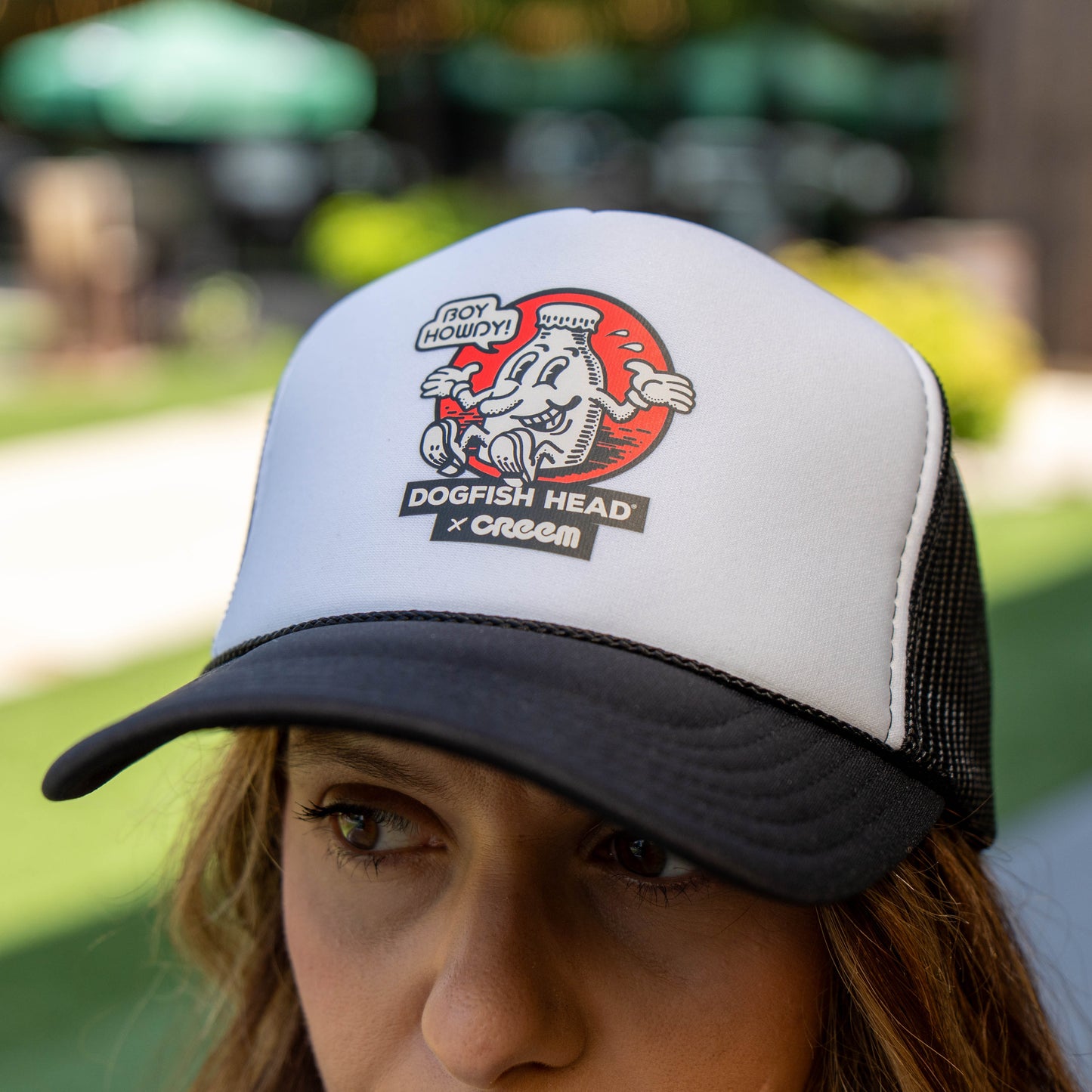 A close-up of a woman wearing a white and black trucker hat with a Dogfish Head x Cream graphic on the front, set against a blurred outdoor patio background.