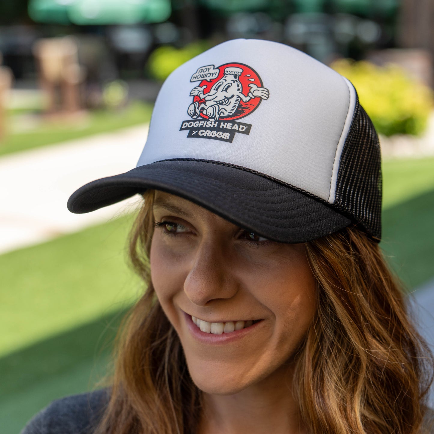 A woman wearing a white and black trucker hat with a 'Dogfish Head &x Creem' graphic on the front, photographed outdoors with a blurred green lawn background.