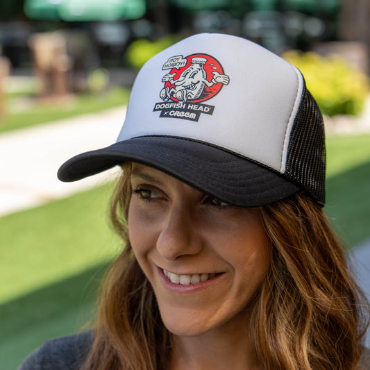 A woman wearing a white and black trucker hat with a 'Dogfish Head &x Creem' graphic on the front, photographed outdoors with a blurred green lawn background.
