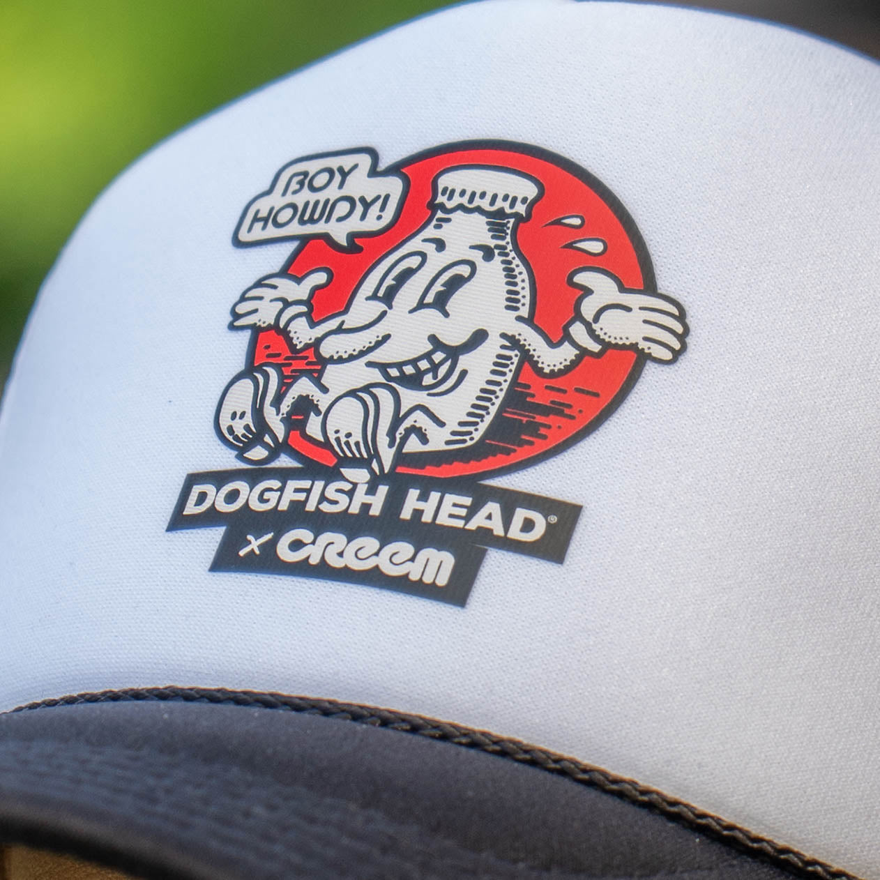 Close-up of a white trucker hat with a black brim, featuring a large embroidered graphic on the front. The graphic shows a dogfish character inside a red circle with the text 'BOY HOWDY!' and 'DOGFISH HEAD x Creem' below.
